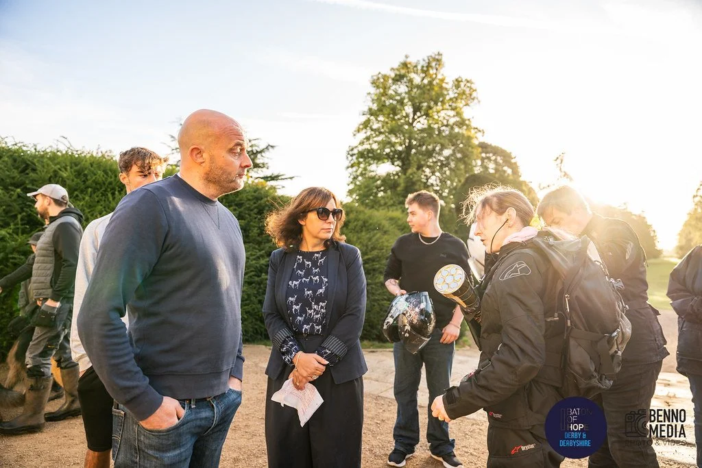 Group of people outdoors, some wearing motorcycle gear, having a discussion during sunset with trees in the background.