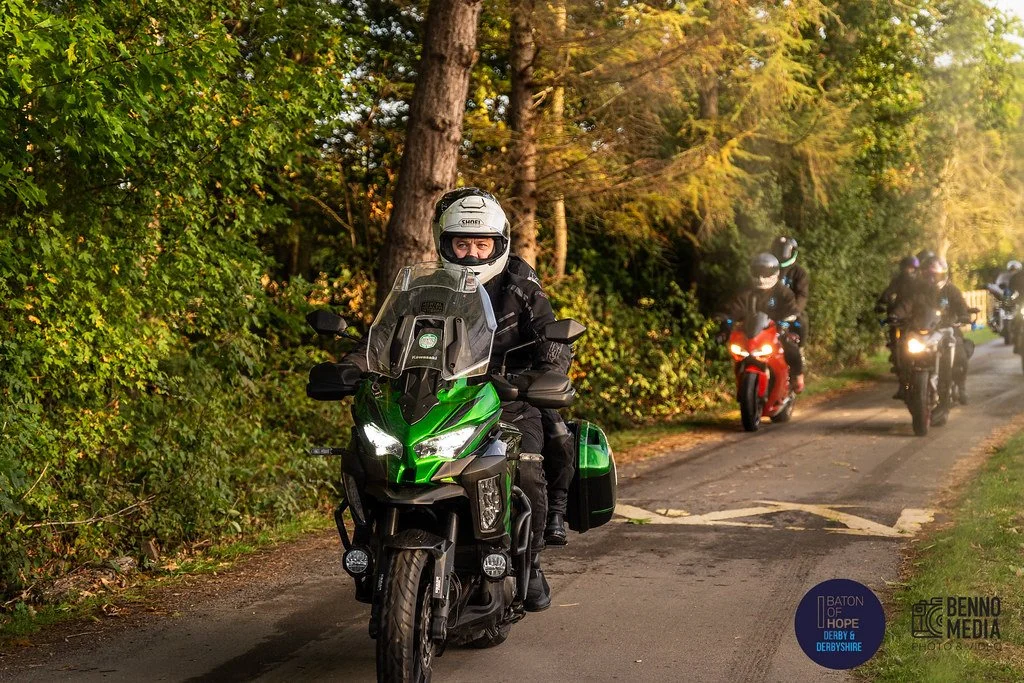 A person riding a green motorcycle on a tree-lined road, with two other motorcyclists riding further back. All riders are wearing helmets and protective gear.