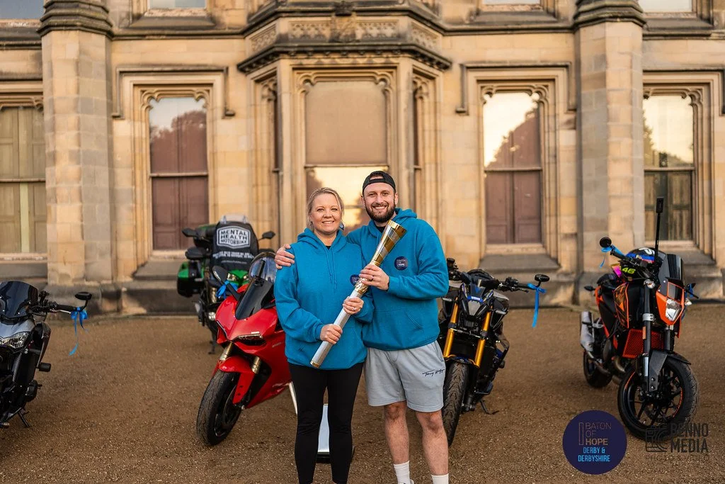 A man and woman standing outdoors in front of a historic building, holding a large torch, with motorcycles parked behind them.