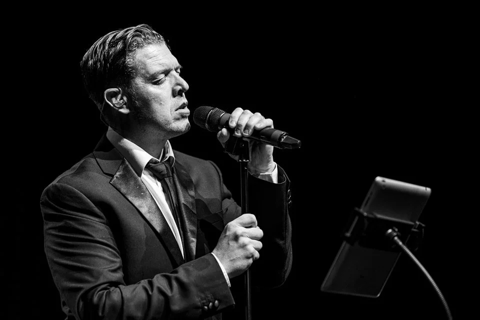 A man in a tuxedo singing into a microphone on stage with a tablet on a stand.
