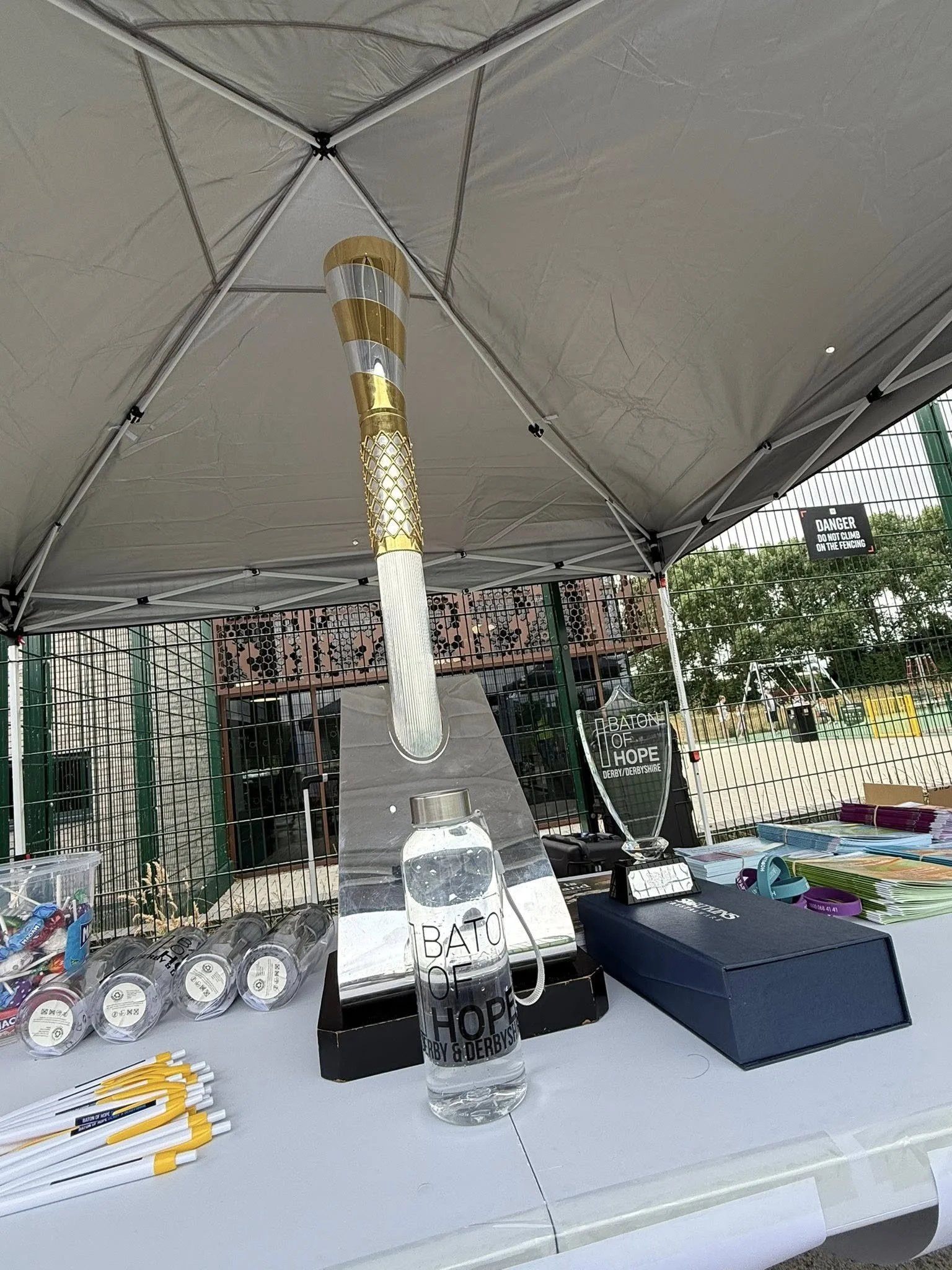 Table at an outdoor event with promotional items, water bottles, and a whistle, with a large baton and a torch in the background, set under a canopy with a fence and trees in the background.