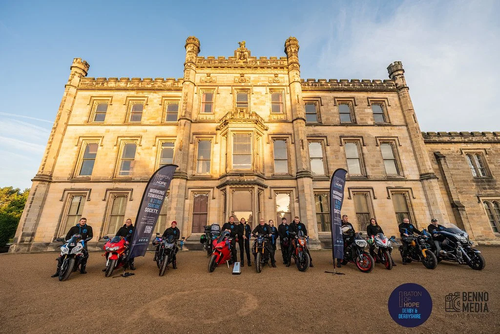 A group of ten people standing next to motorcycles in front of a historic castle with banners that read 'Baton of Hope.'