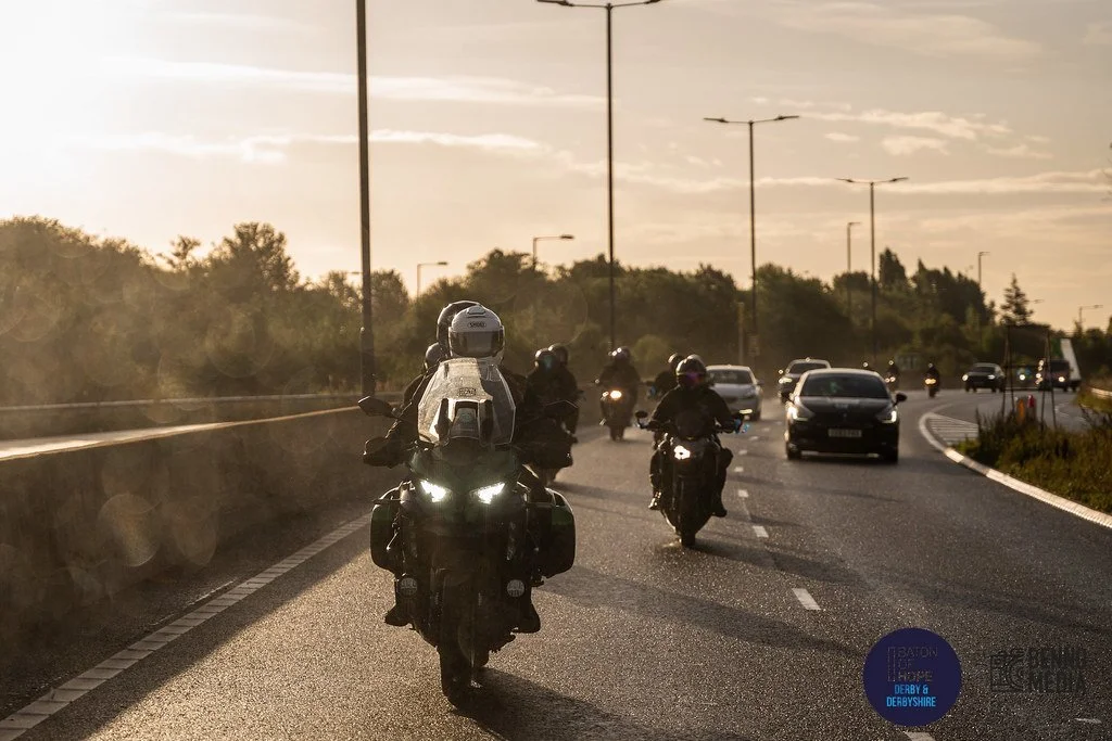 Motorcycles and cars traveling on a highway during the daytime, with trees on the side and street lights above.