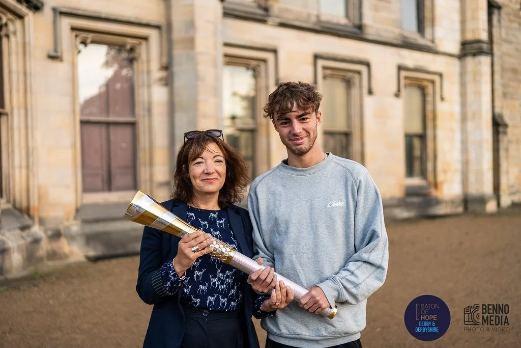 A woman and a young man standing outside on a dirt ground, holding a rolled-up poster or banner, smiling at the camera. The woman has glasses on her head and is wearing a dark blue jacket, while the young man is dressed in a light gray sweatshirt. Th