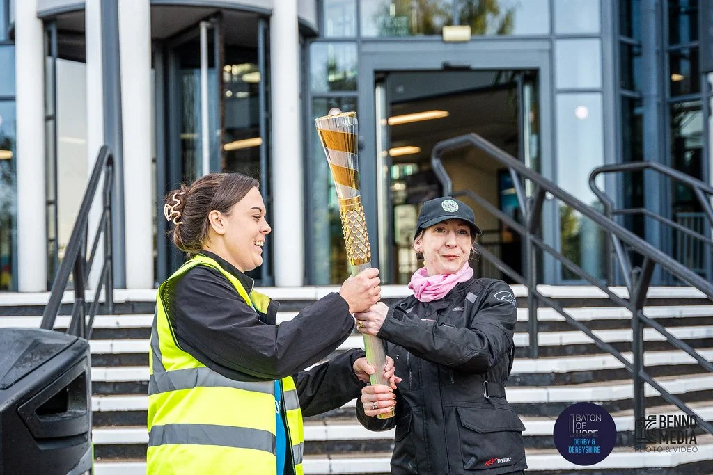 Two women, one in a yellow reflective vest and the other in a black jacket and cap, holding a torch together outside a building with stairs, celebrating a sporting event.