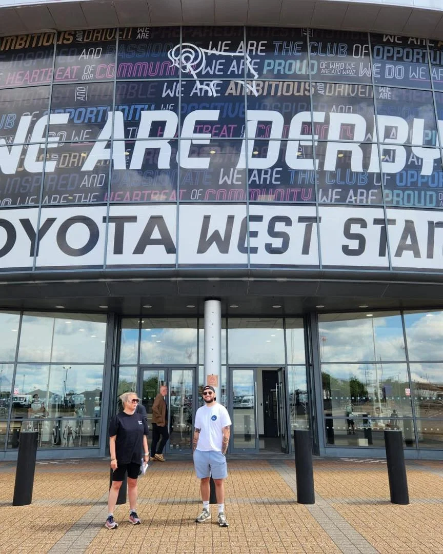 People standing outside Toyota West Start dealership in front of a large glass building with a sign reading "WE ARE DRAFT" and development-related words on the windows.