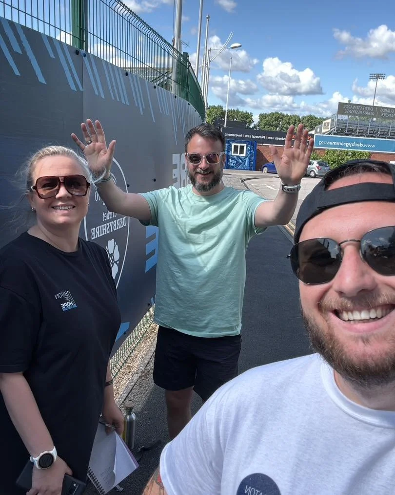Three smiling people taking a selfie outdoors near a sports field, with a large sign and a fence behind them, on a sunny day with a partly cloudy sky.