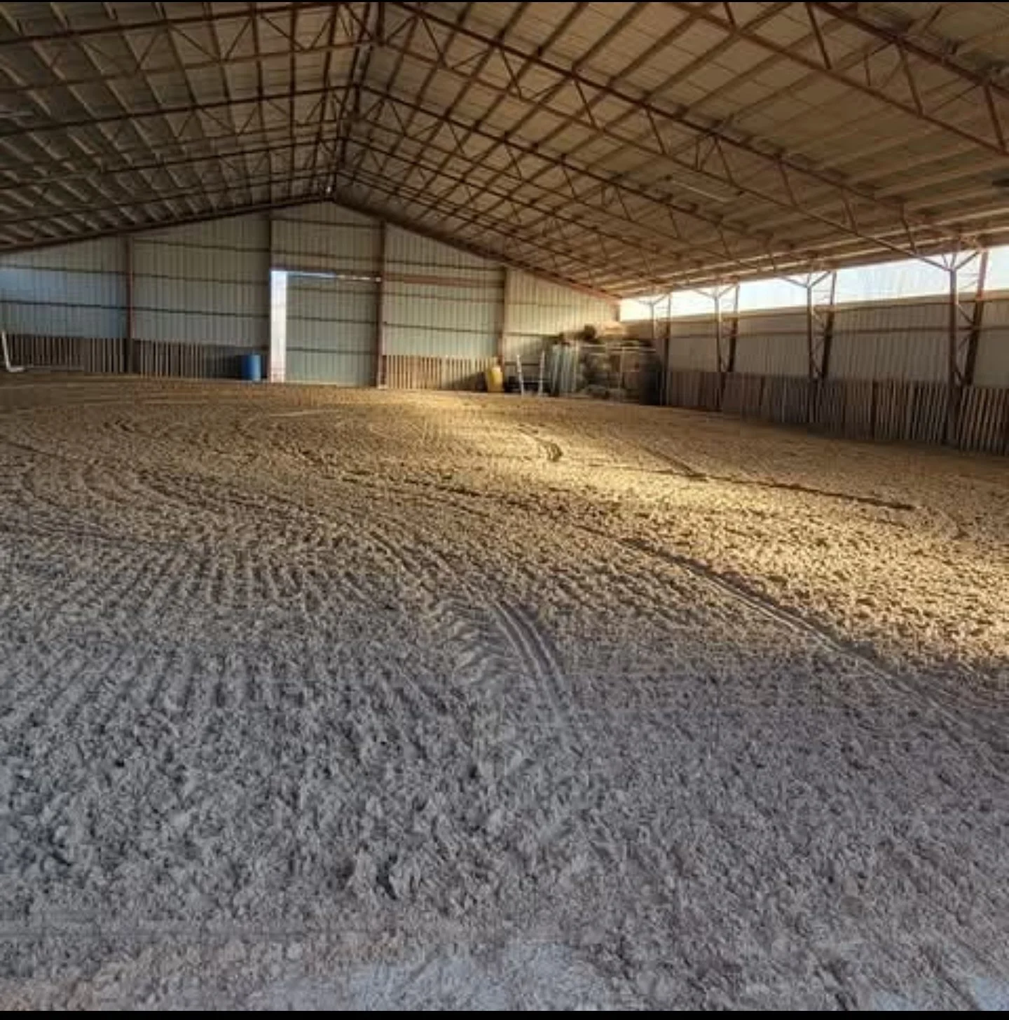 Empty indoor equestrian arena with a dirt or sand riding surface and metal walls, illuminated by natural light.