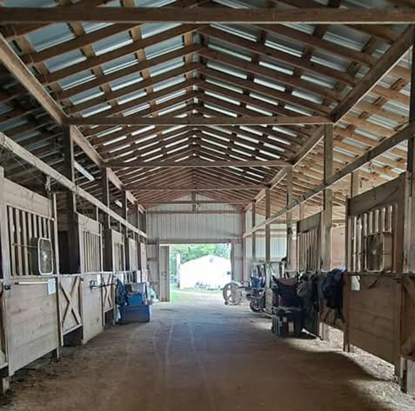 Interior of a stable or barn with wooden stalls on either side, hay or dirt floor, and an open door at the end letting in natural light.
