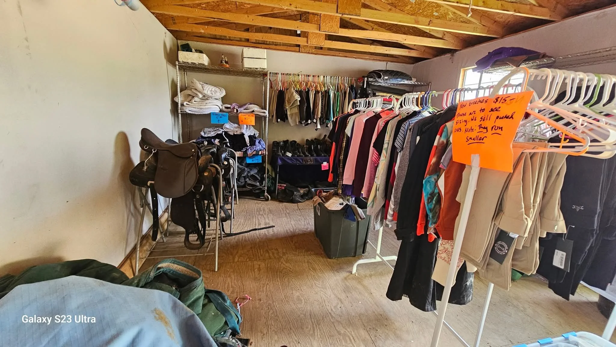 A small indoor thrift shop with clothing racks, shelves, and miscellaneous items. The room has a wooden ceiling and a window on the right, with a mix of hanging clothes and folded items. A bright orange sign indicates prices and sales information.