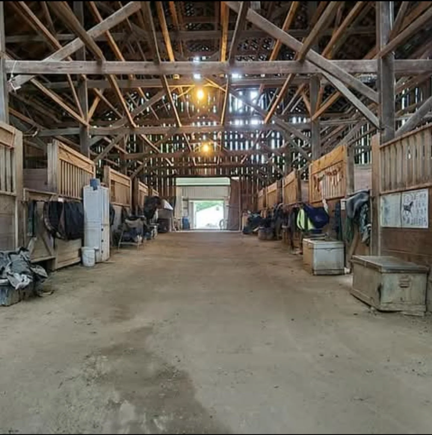 Indoor barn with wooden walls and ceiling, showing equipment, bags, and boxes along the sides, with a dirt floor and open doors letting in natural light.