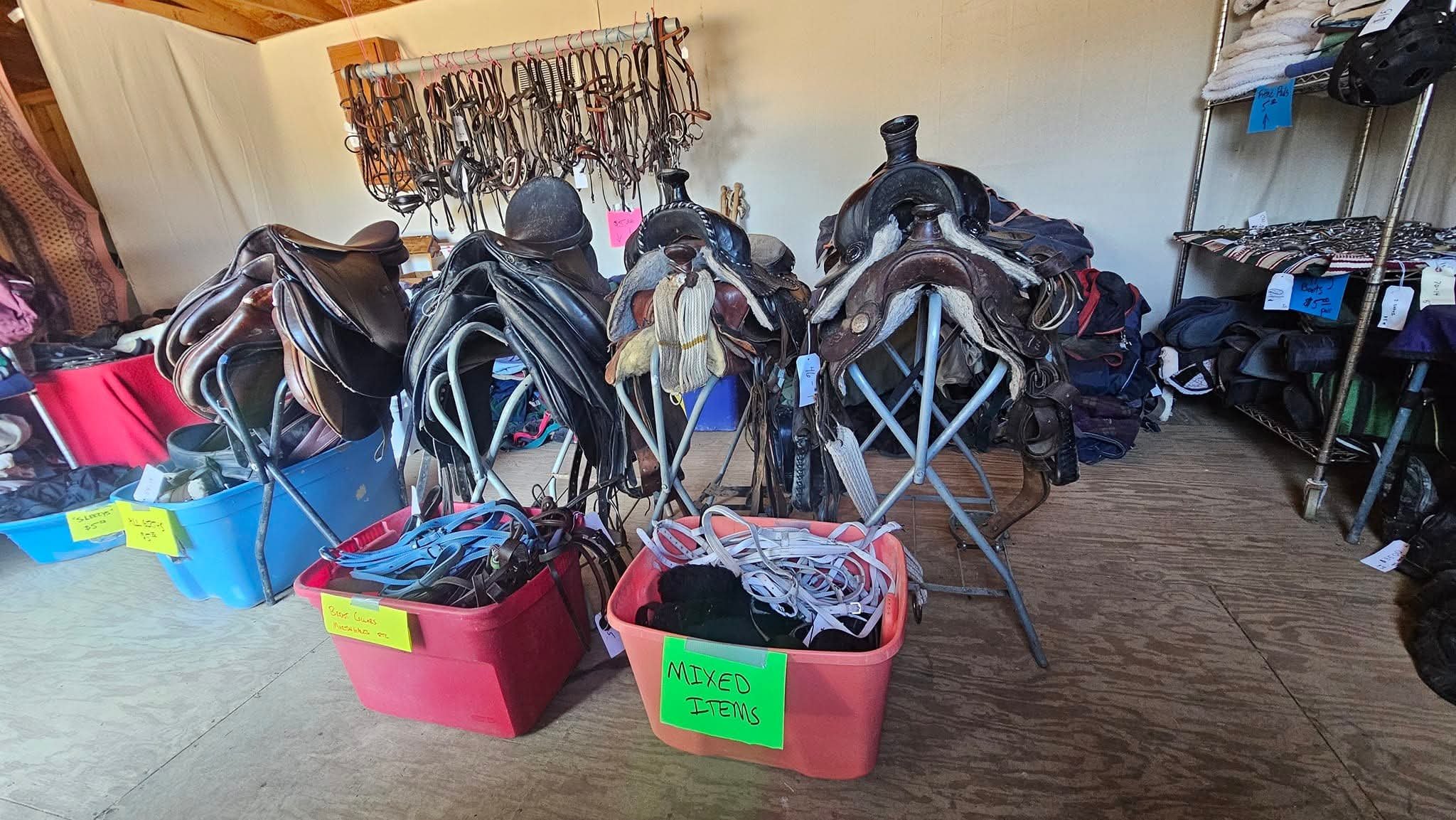 Display of used horse riding equipment including saddles, bridles, stirrups, and helmets arranged on tables and in bins at a resale store.