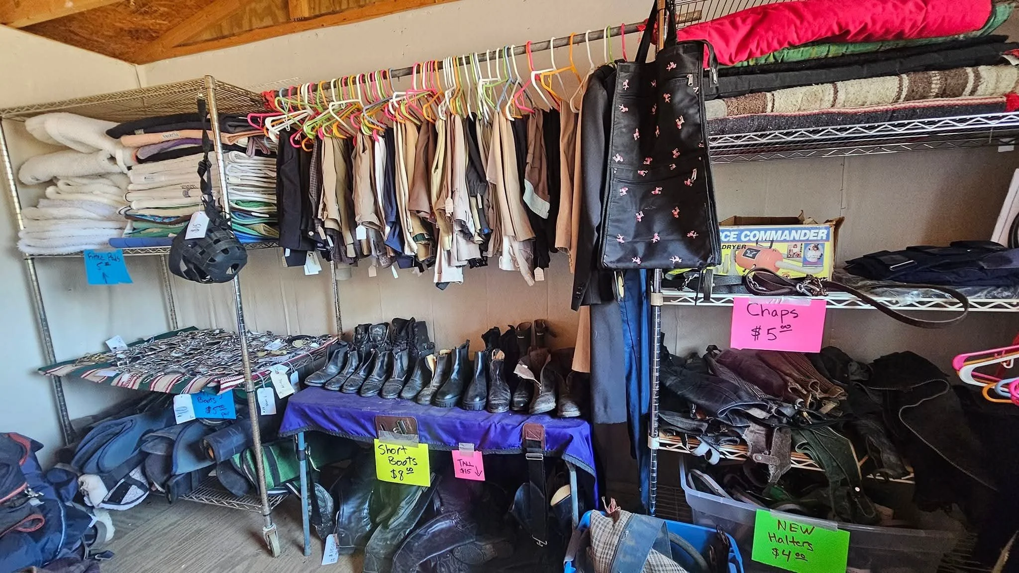 A thrift store display with clothing, shoes, and accessories. Shelves hold folded towels, and racks feature hanging shirts, pants, and jackets. Signage indicates prices for chaps, boots, and hats.