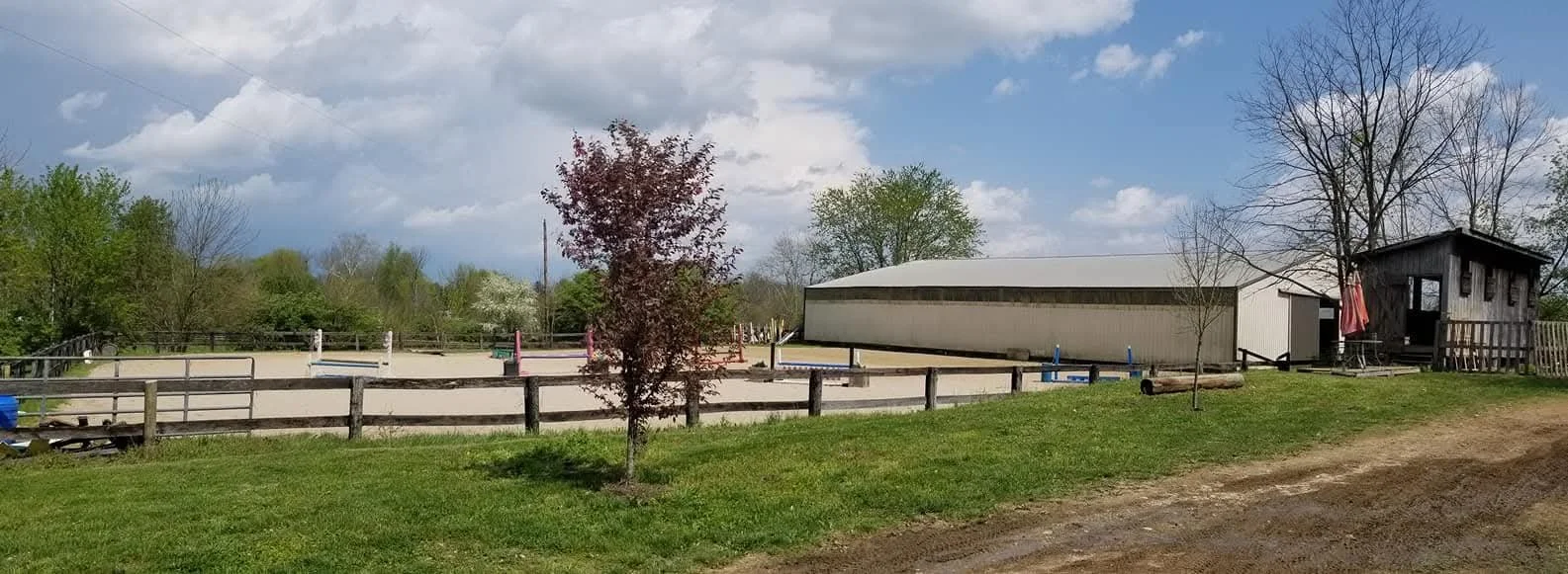An outdoor equestrian riding arena with a wooden fence, surrounded by trees and a small wooden building, under a partly cloudy sky.