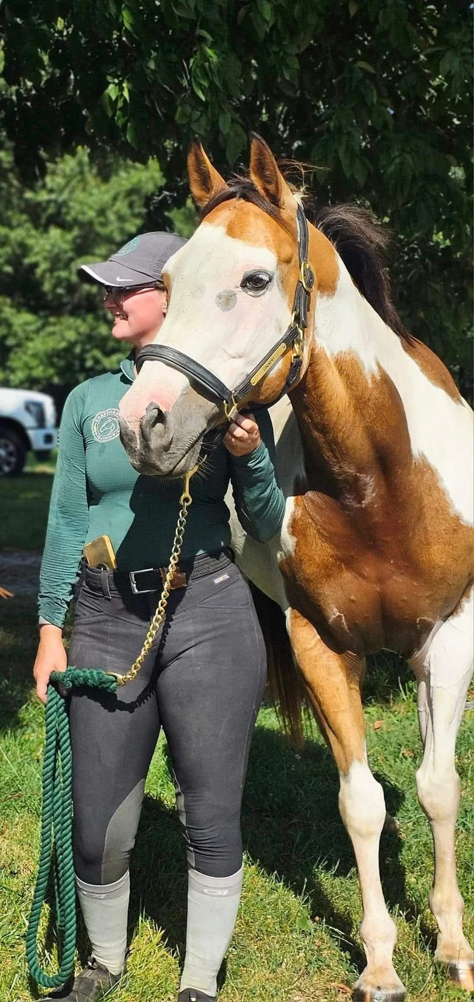 A woman in riding gear holding a rope leads a large horse with a distinctive white and brown coat, standing outdoors with green foliage and a vehicle in the background.