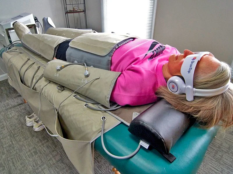 Person in FlowPresso compression suit lying on a massage table outdoors, surrounded by greenery, wearing eye mask and headphones.