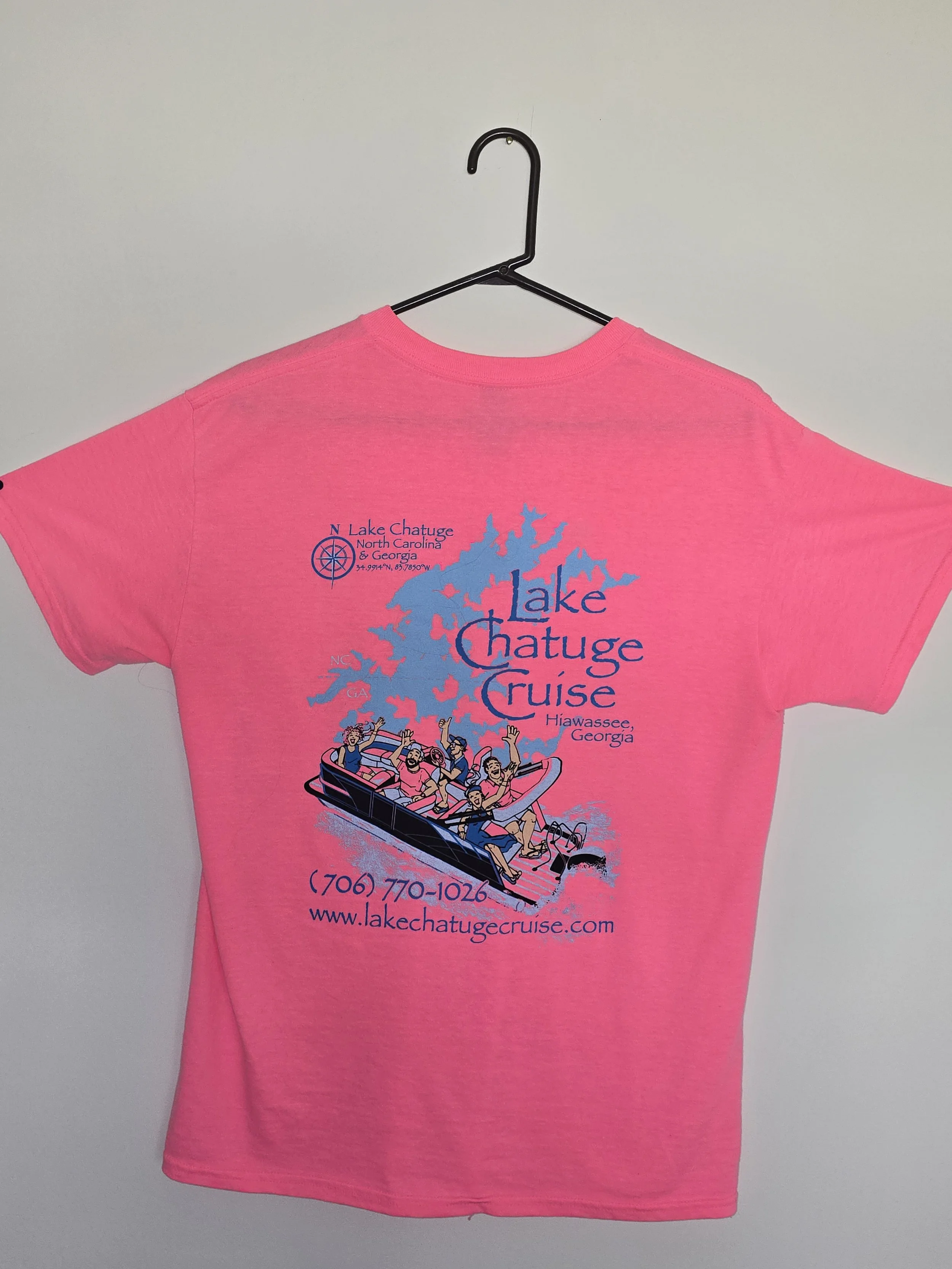 Pink T-shirt with graphic and text, attached to a black hanger, promoting Lake Chatuge Cruise in Georgia, with a cartoon of people on a boat, a map outline, web address, and contact number.