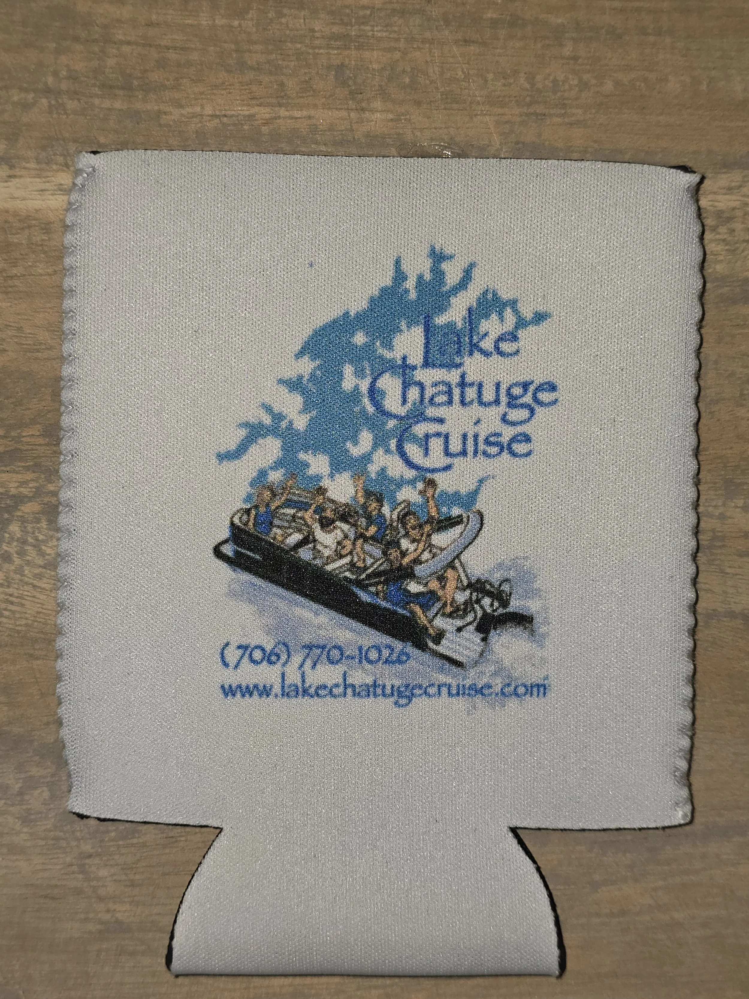 A souvenir fabric drink koozie with a motel logo for Lake Chatugue Cruise, featuring an illustration of a boat with people tubing behind it, a blue pine tree, and contact information.