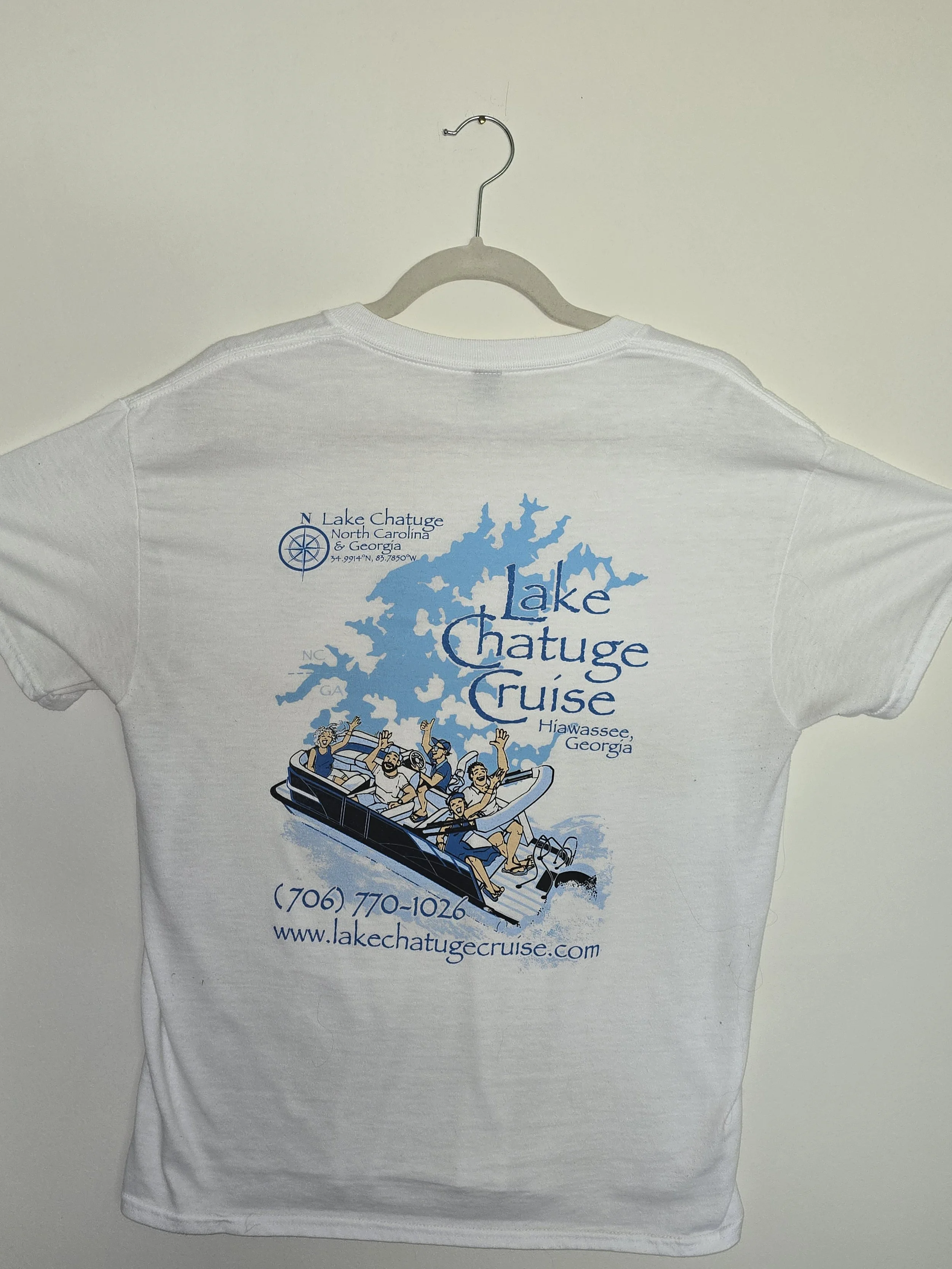 White T-shirt hanging on a hanger with a colorful graphic design of a lakeside scene, including a boat with people waving, and text promoting Lake Chatuge cruise in Georgia with a website and phone number.