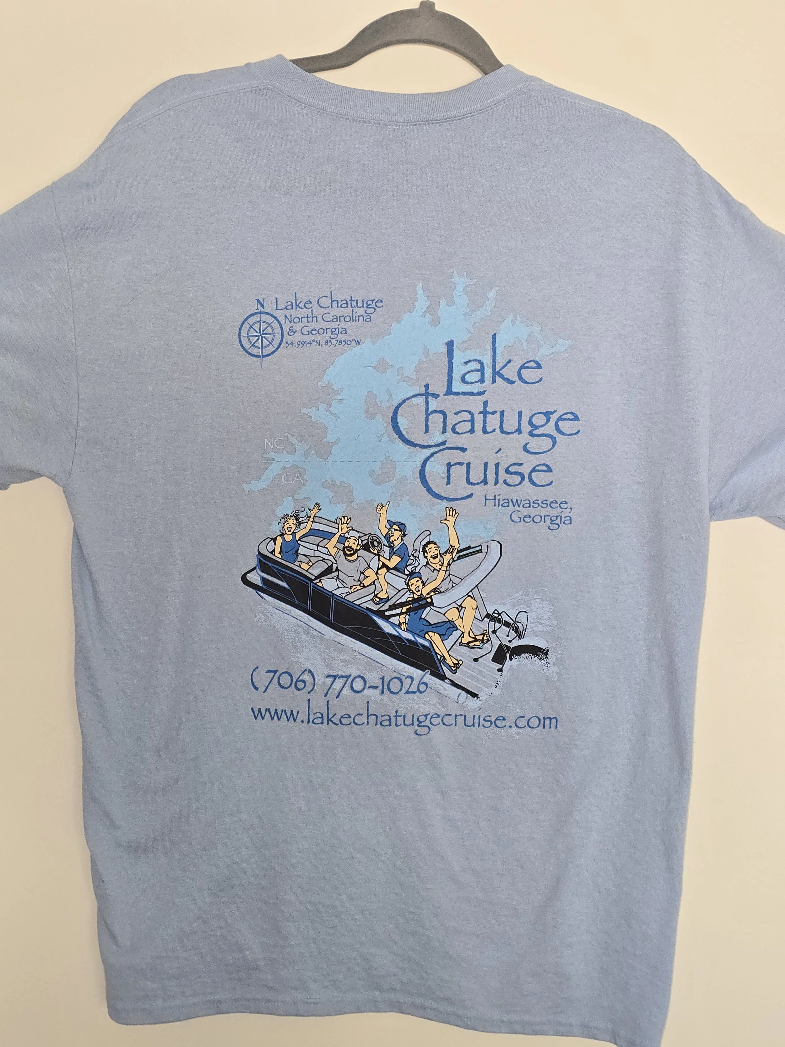 Light blue T-shirt with a graphic design promoting Lake Chatuge Cruise in Hiawassee, Georgia, featuring a cartoon boat with six people waving, along with a map outline and contact information.
