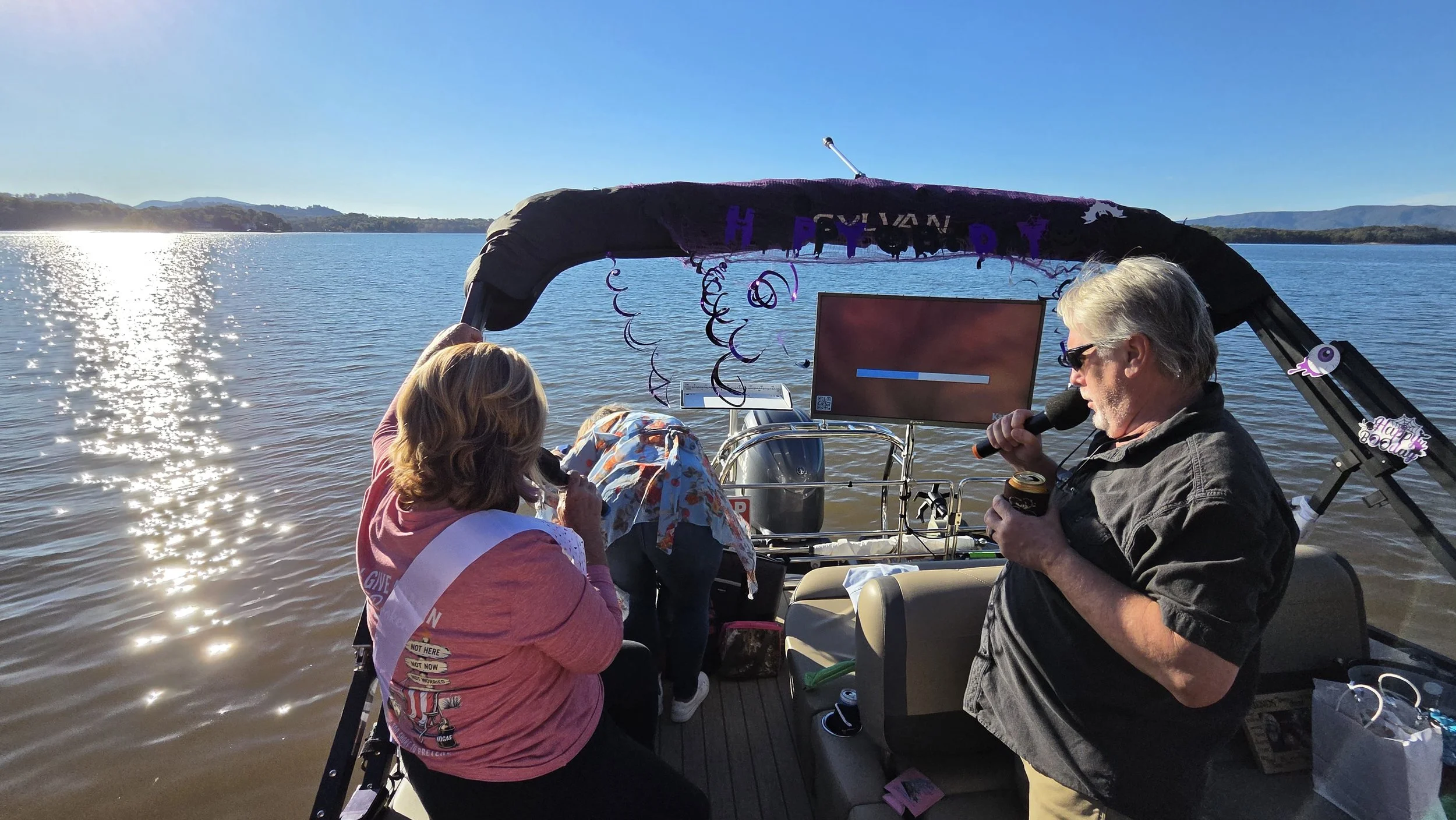 A lively scene on a boat during sunset, with people singing, dancing, and celebrating at a karaoke cruise on Lake Chatuge. Bright neon signs promote karaoke, boat tours, bachelorette parties, and corporate gatherings, creating an energetic party atmosphere.