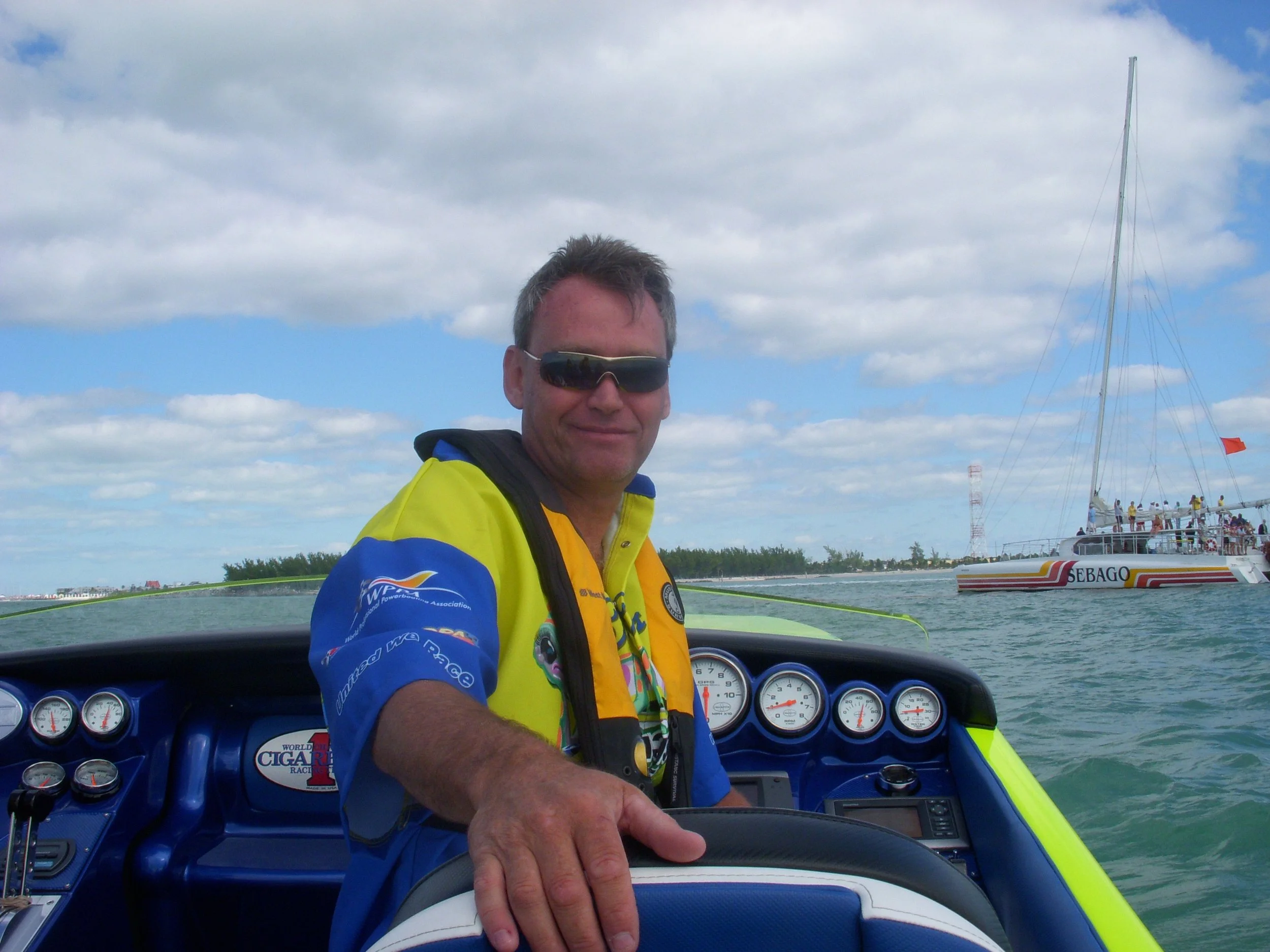 Man on speedboat with colorful gauges, wearing sunglasses and a bright jacket, ocean and sailboat in background under cloudy sky.