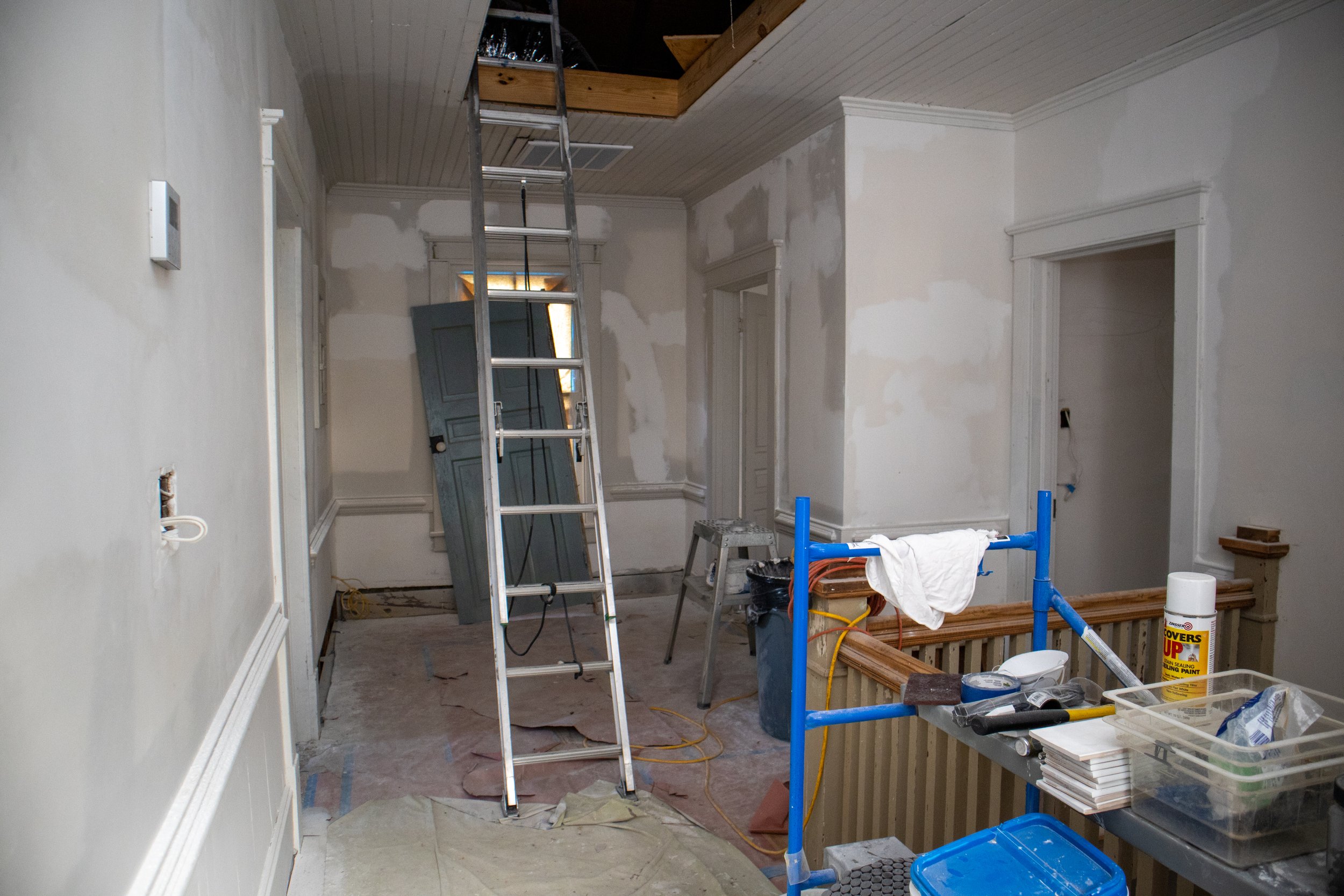 Room under renovation with ladder leading to attic, painting supplies, drywall patches, and a door leaning against the wall.