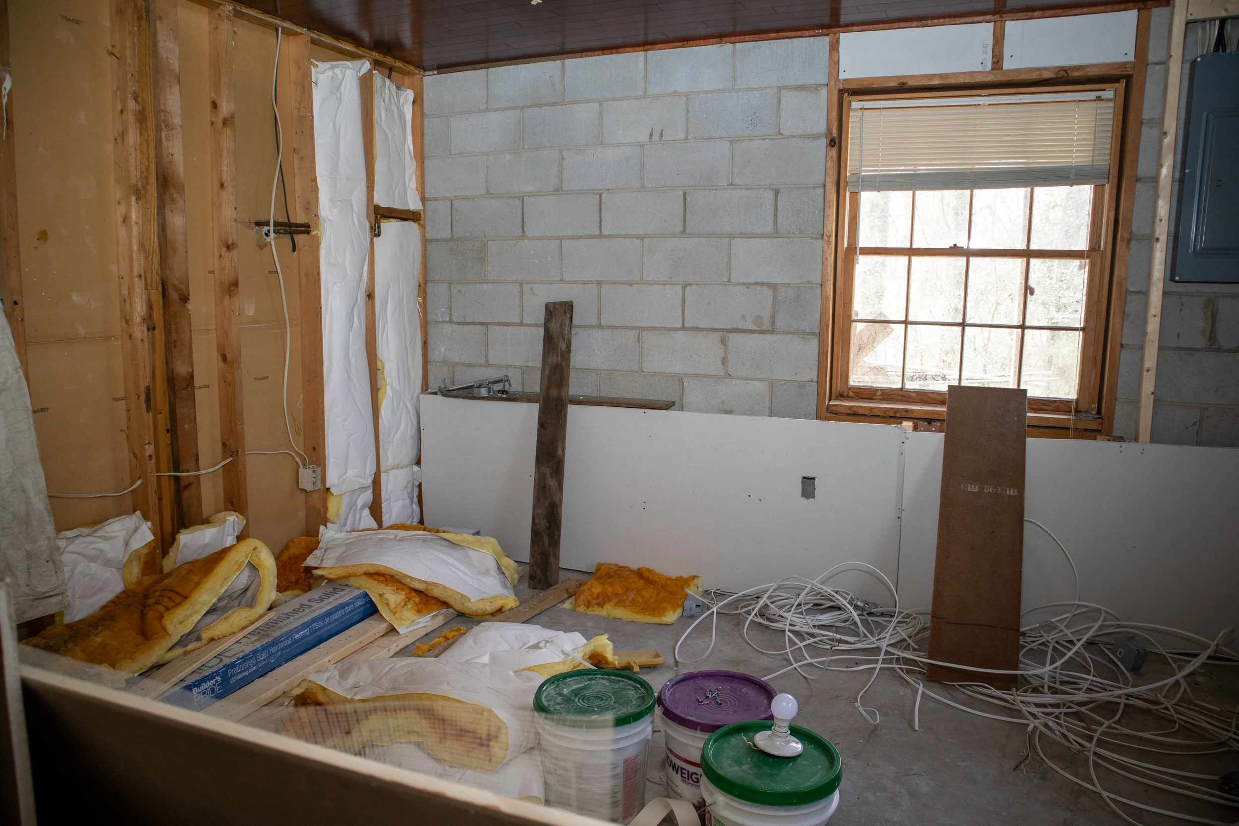 Room under construction with exposed insulation, stacked drywall, electrical cords, window, and paint buckets.