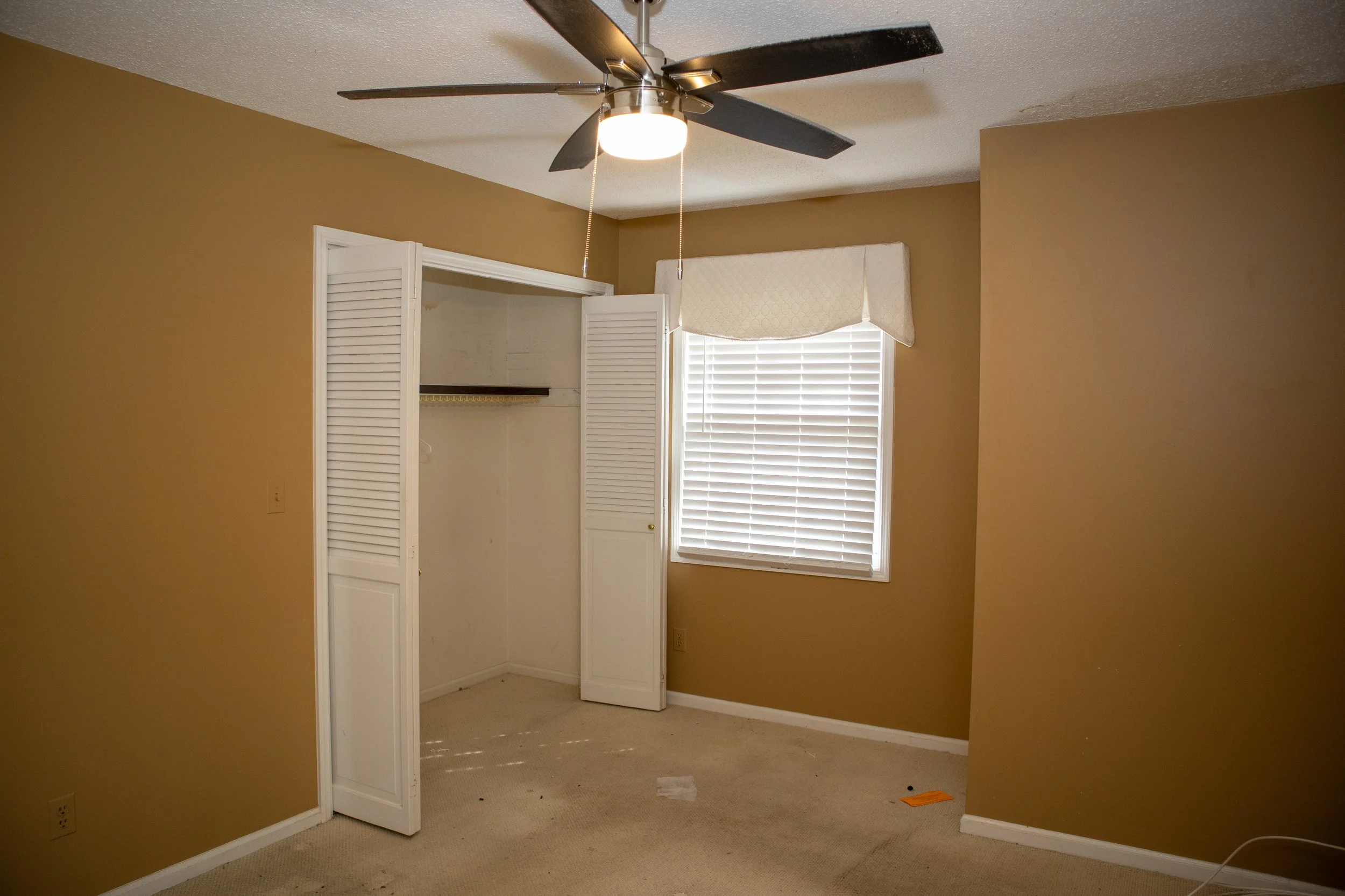 Empty room with a ceiling fan, beige walls, an open closet with white doors, and a window with blinds. The floor is carpeted.