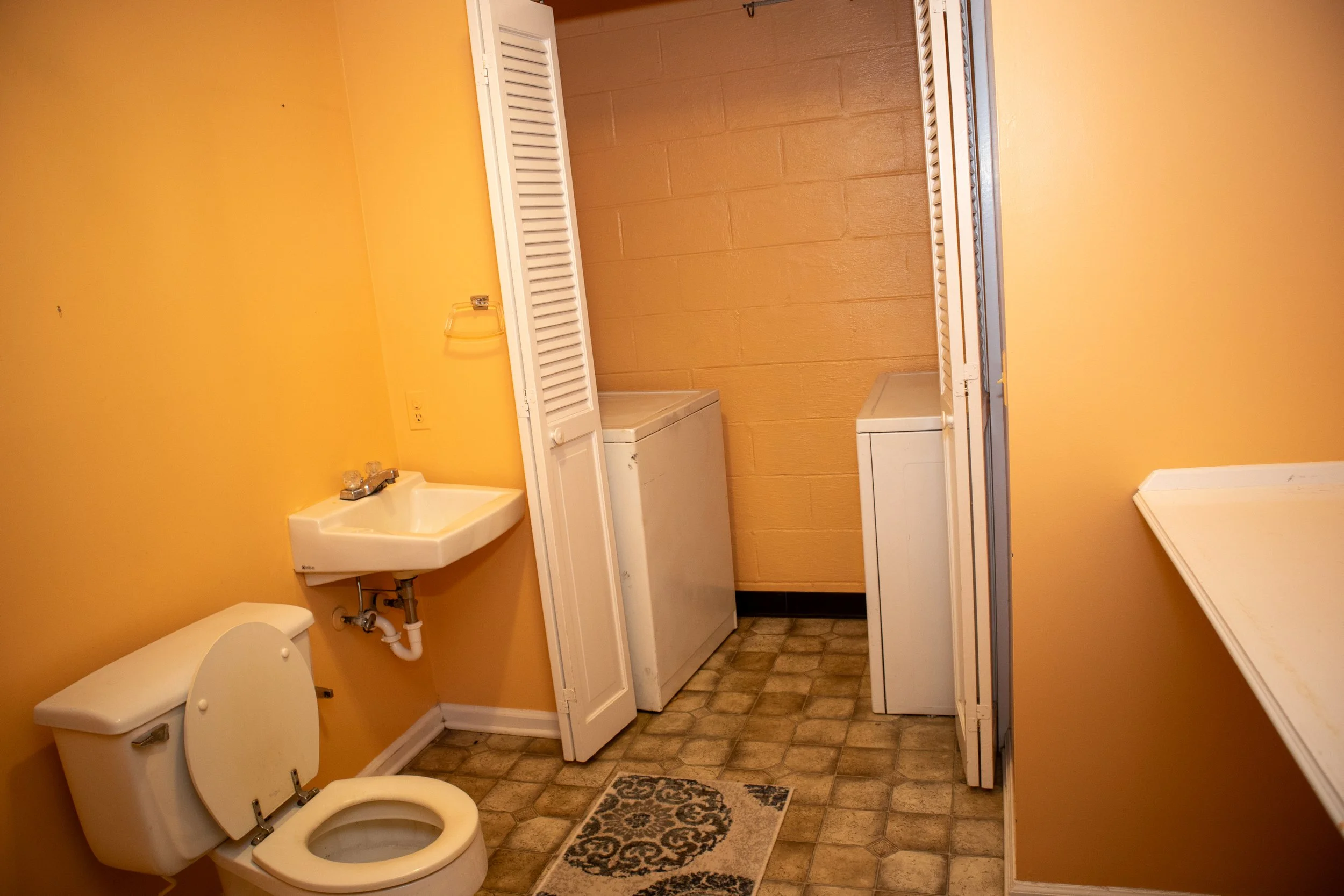 Small bathroom with a toilet, sink, washing machine, and dryer, featuring orange walls and tiled floor.