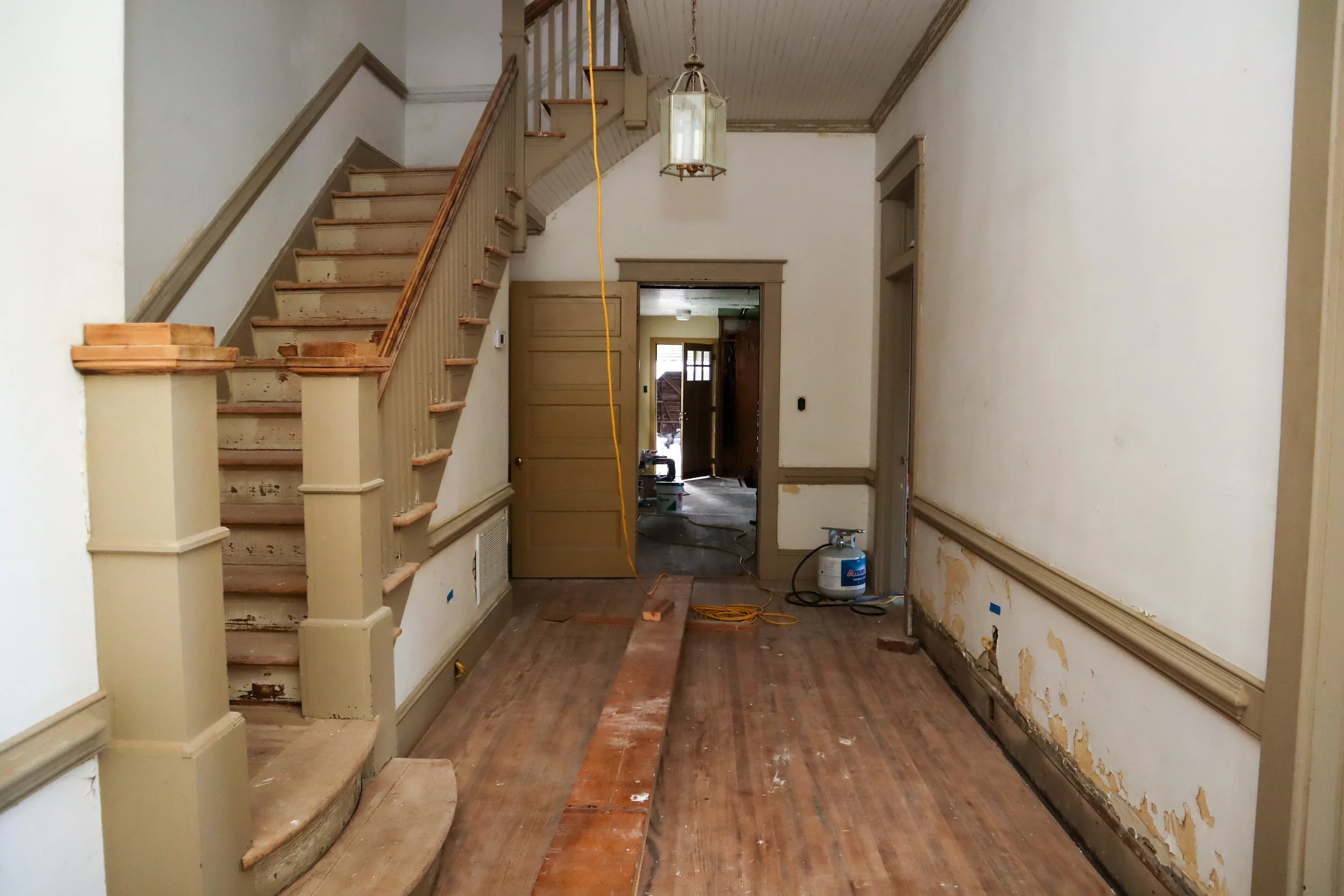 Old house hallway under renovation with exposed wooden floors, peeling paint, a staircase on the left, and a light fixture hanging from the ceiling.