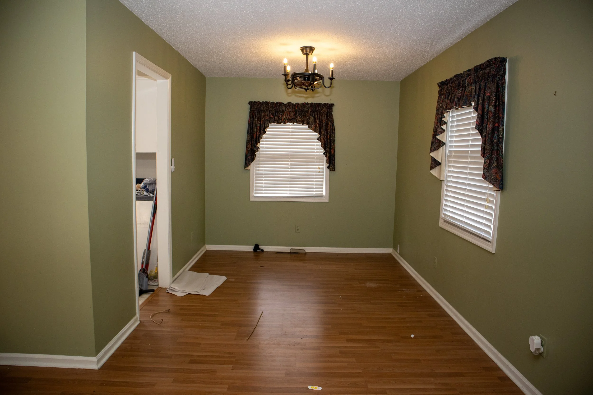 Empty room with green walls, wooden floor, two windows with dark curtains, a ceiling light fixture, and a doorway leading to another room. Cleaning supplies are visible near the doorway.