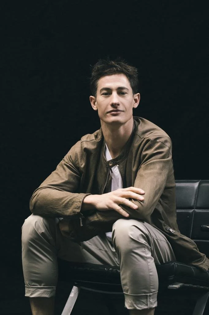 A young man with short dark hair, wearing a brown jacket and beige pants, sitting on a black chair against a dark background, looking at the camera with a neutral expression.
