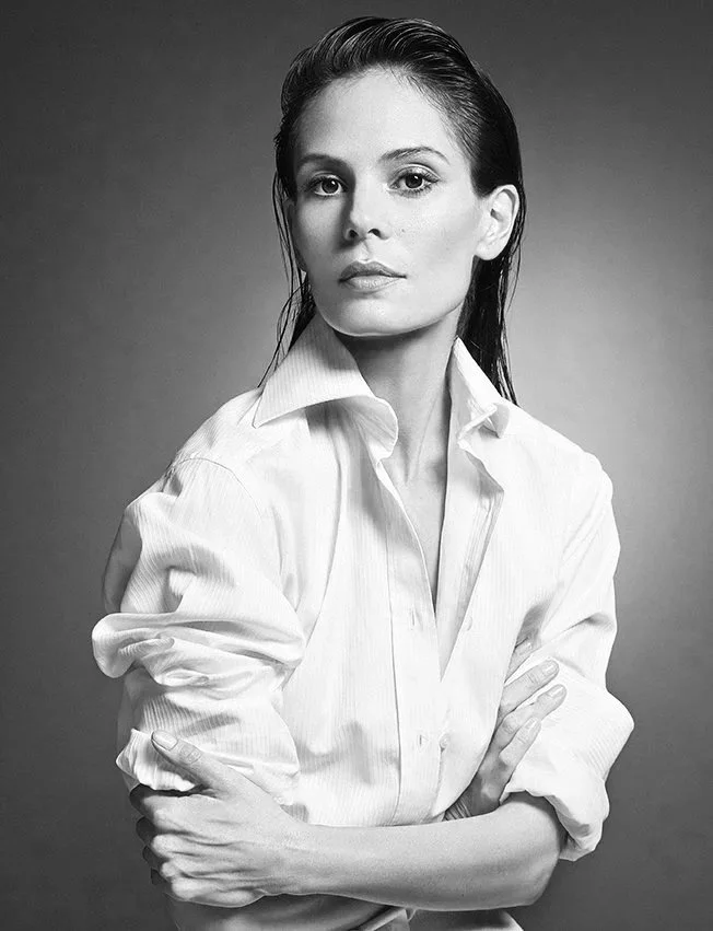 Black and white portrait of a woman with short, slicked-back hair wearing a white button-down shirt with rolled-up sleeves, crossing her arms, looking confidently at the camera.
