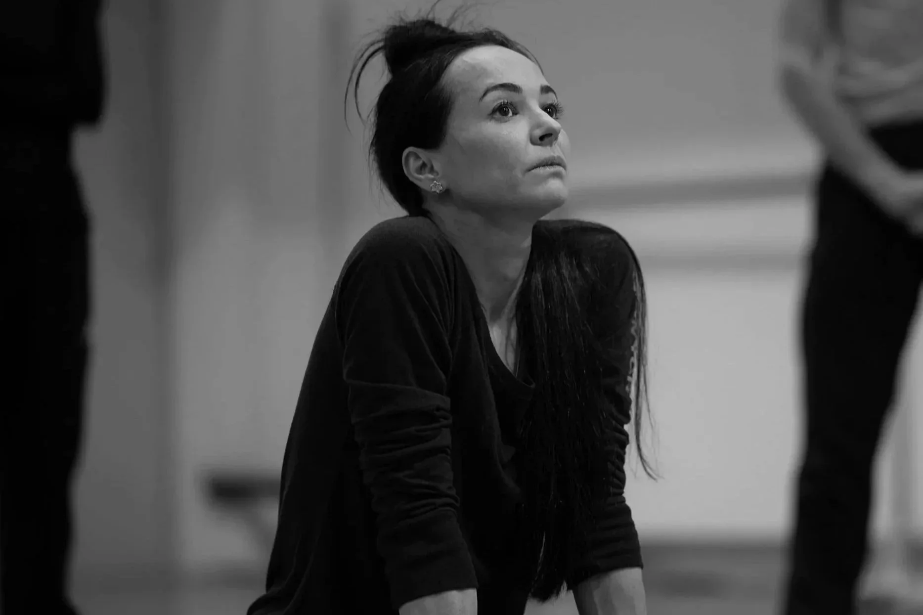 A woman with dark, wet hair, wearing earrings and an intense expression, is seated on the floor in a black dress, looking upwards, in a monochrome setting.