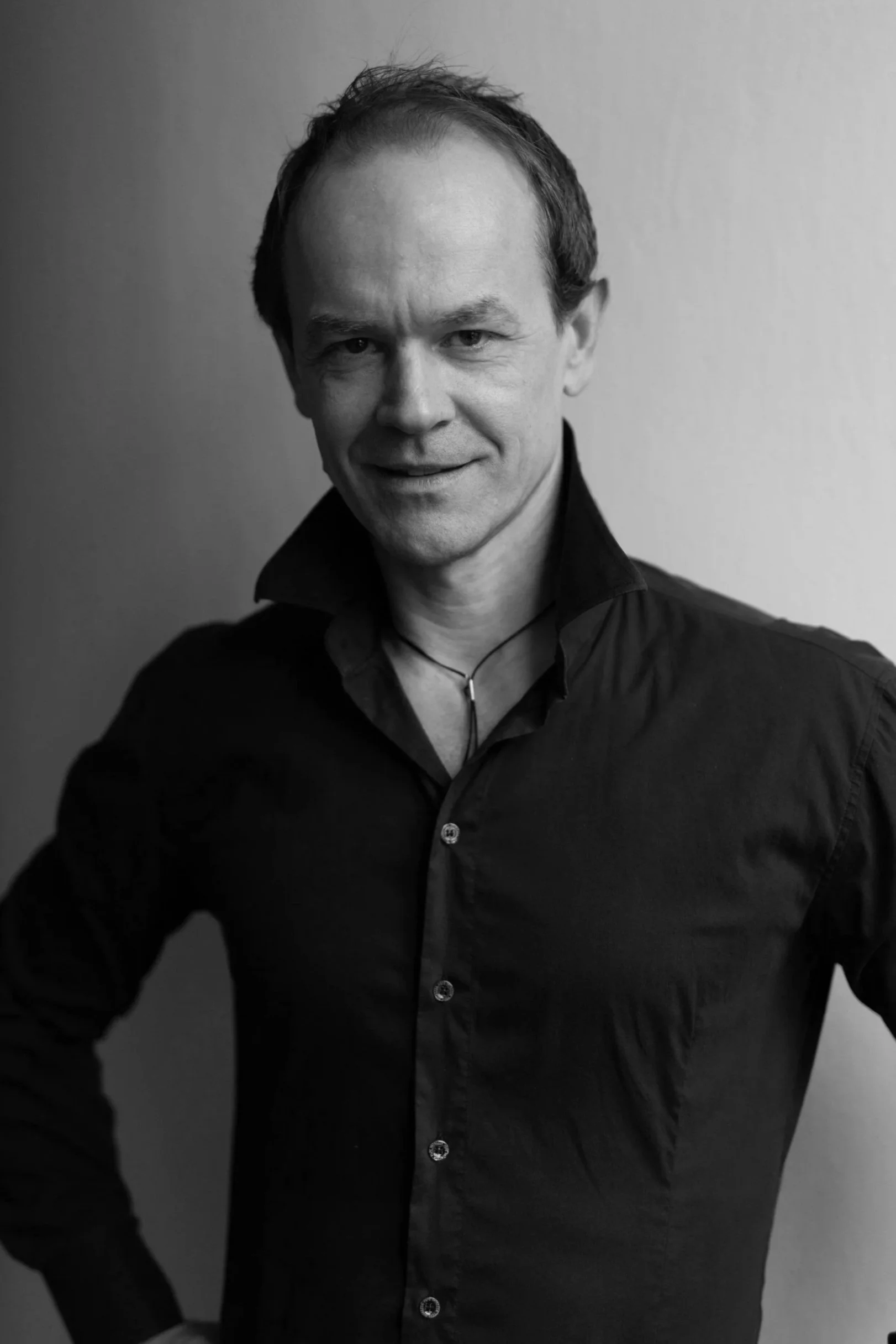 Black and white portrait of a man with short hair, wearing a collared shirt and a necklace, standing against a plain background.