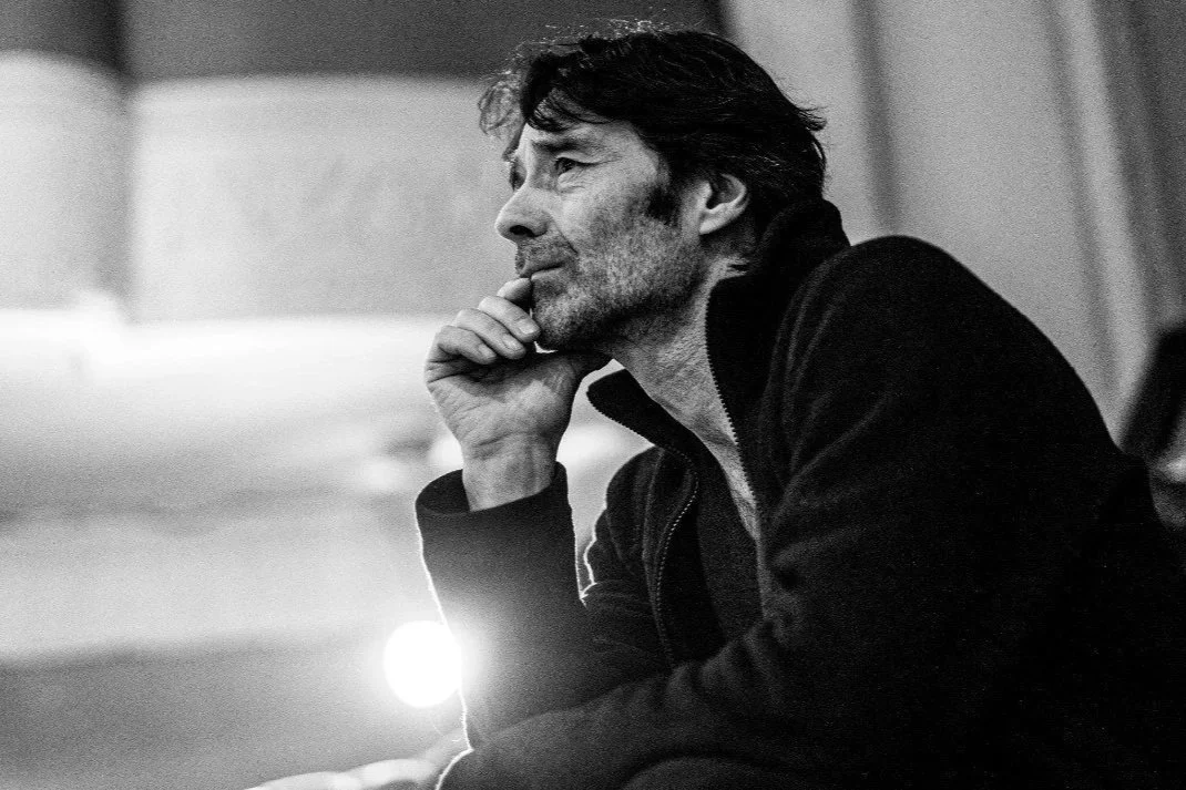 A black and white photo of a man with dark hair and a beard, sitting and resting his chin on his hand, looking thoughtfully.