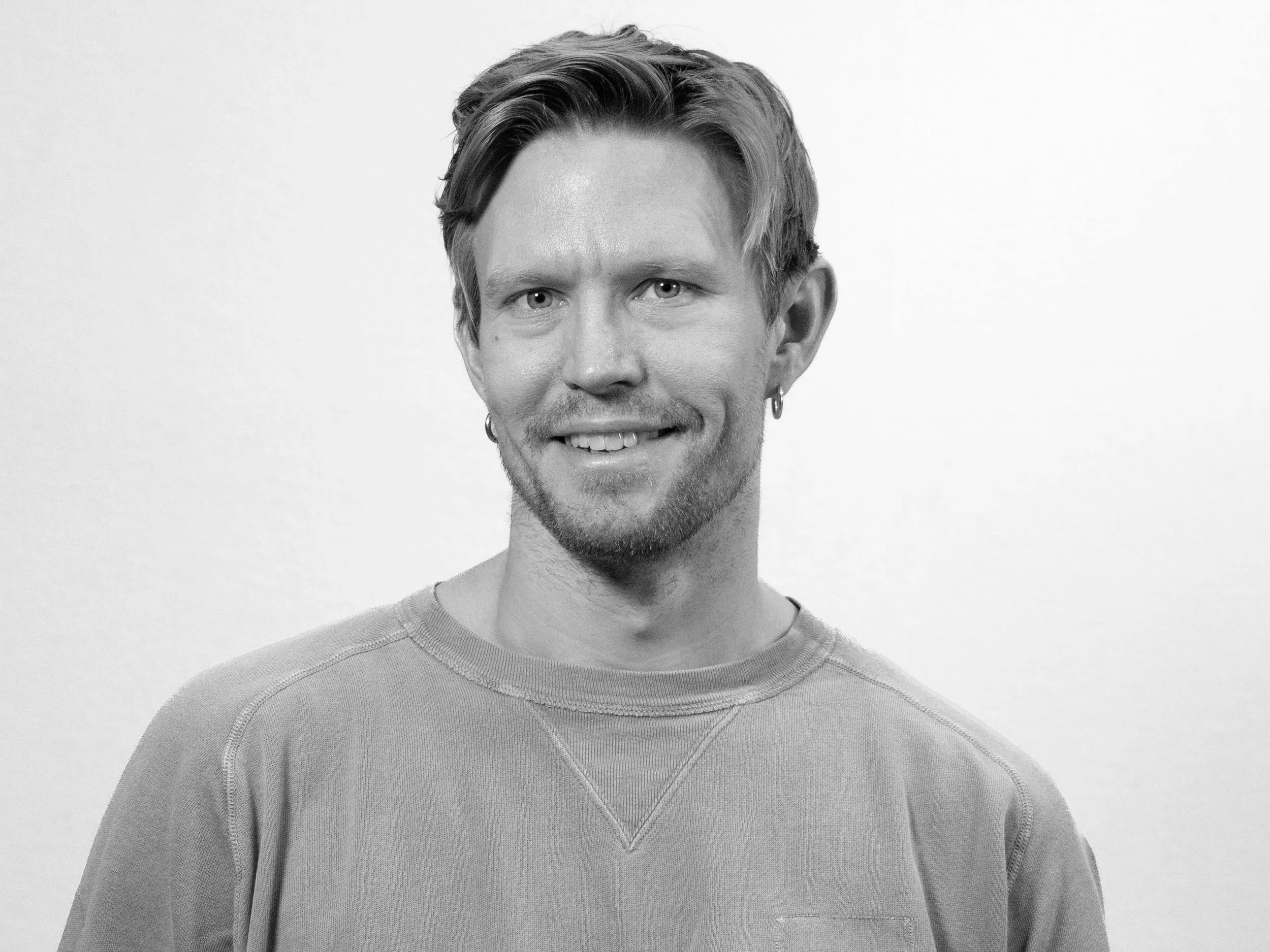 Black and white portrait of a smiling young man with light hair and earrings, wearing a sweatshirt, standing against a plain background.