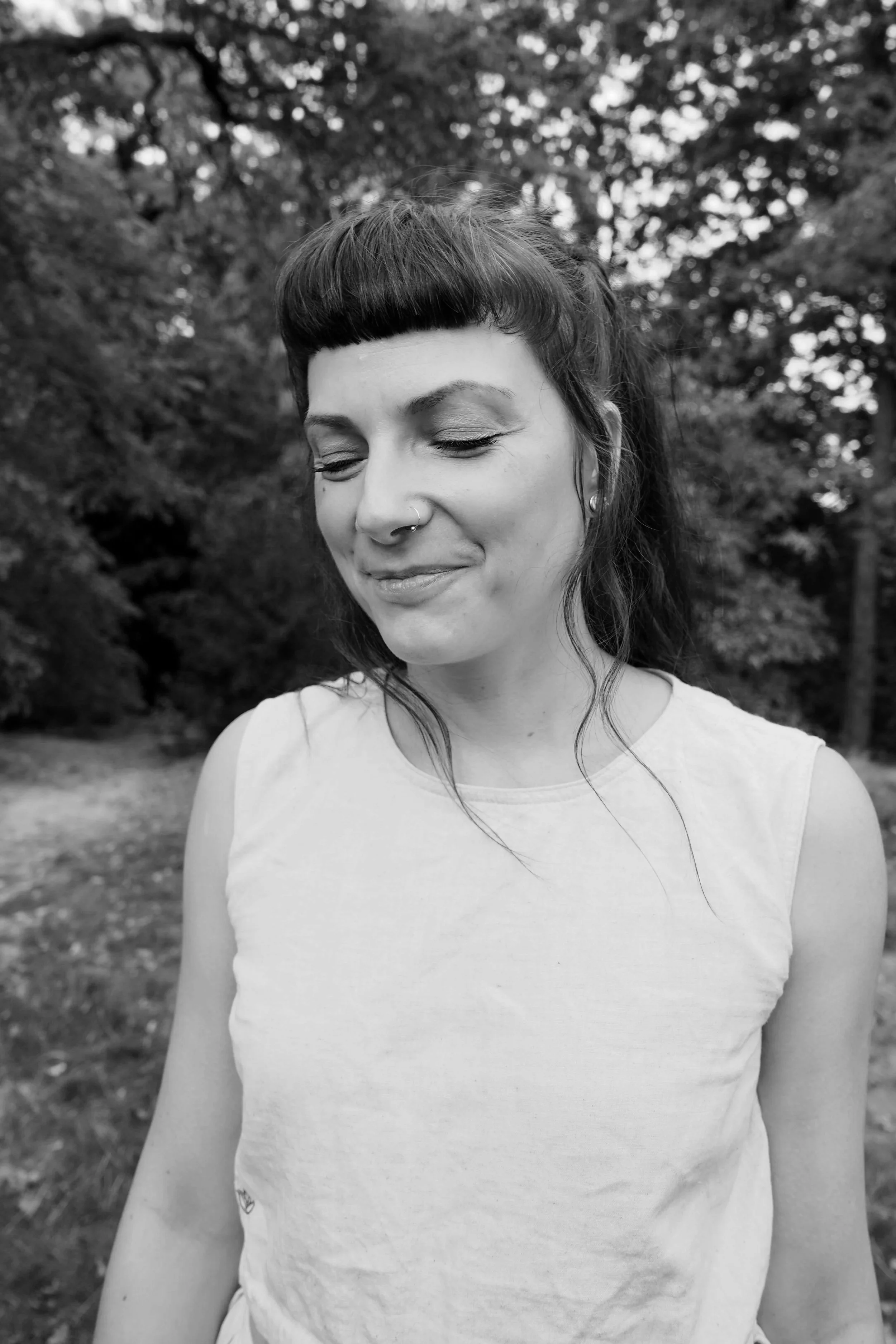 A woman with dark hair and bangs, closed eyes, and a slight smile, standing outdoors with trees in the background.