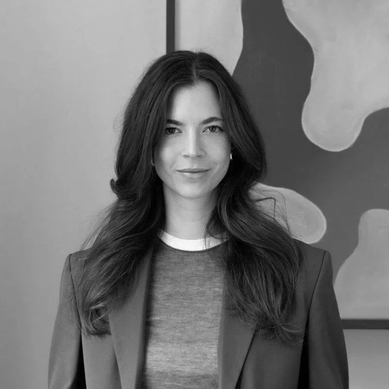 A black-and-white portrait of a woman with long, wavy hair, wearing a blazer over a t-shirt, standing in front of an abstract art piece.