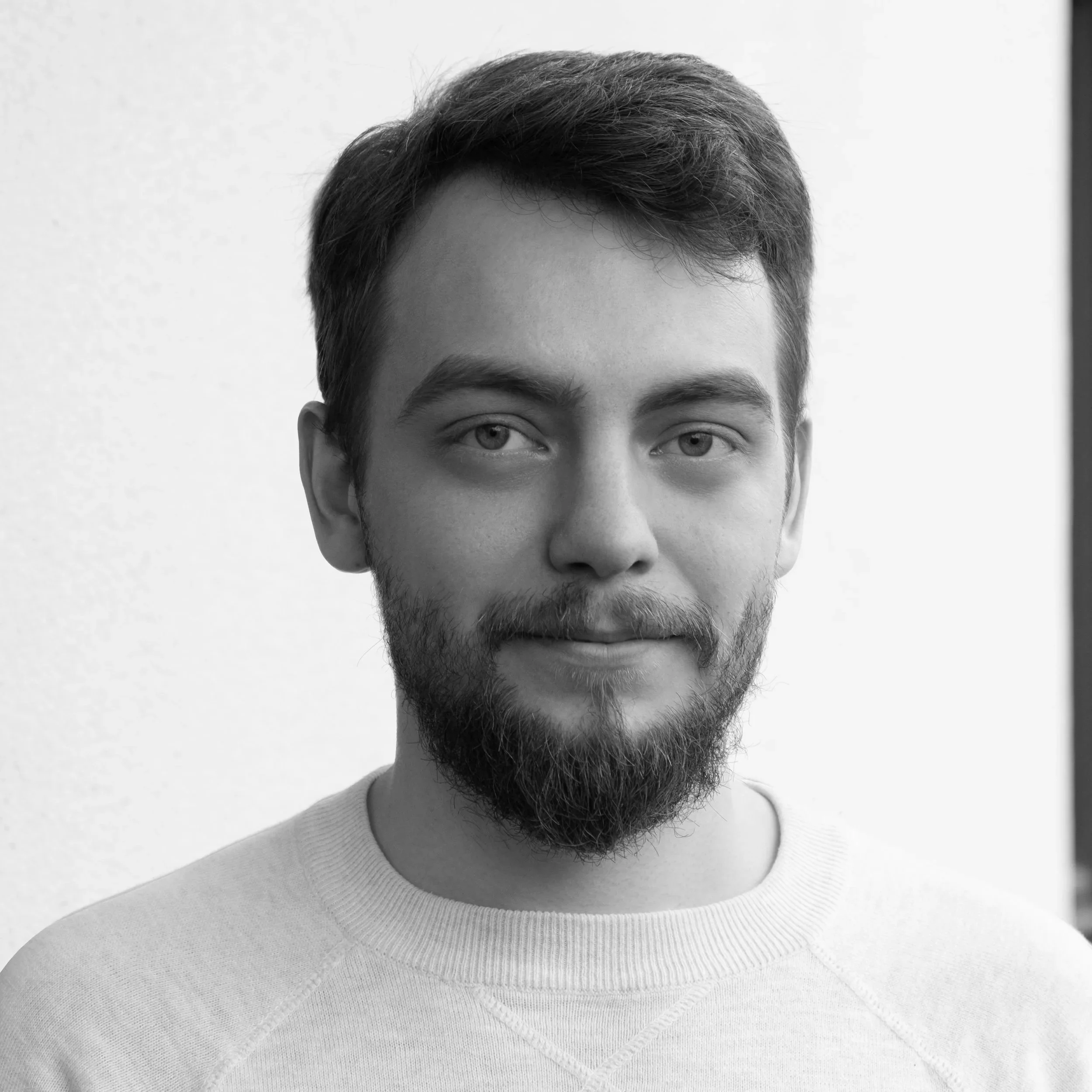 Black and white portrait of a young man with a full beard, short hair, and light-colored eyes, wearing a light-colored shirt, facing the camera with a neutral background.