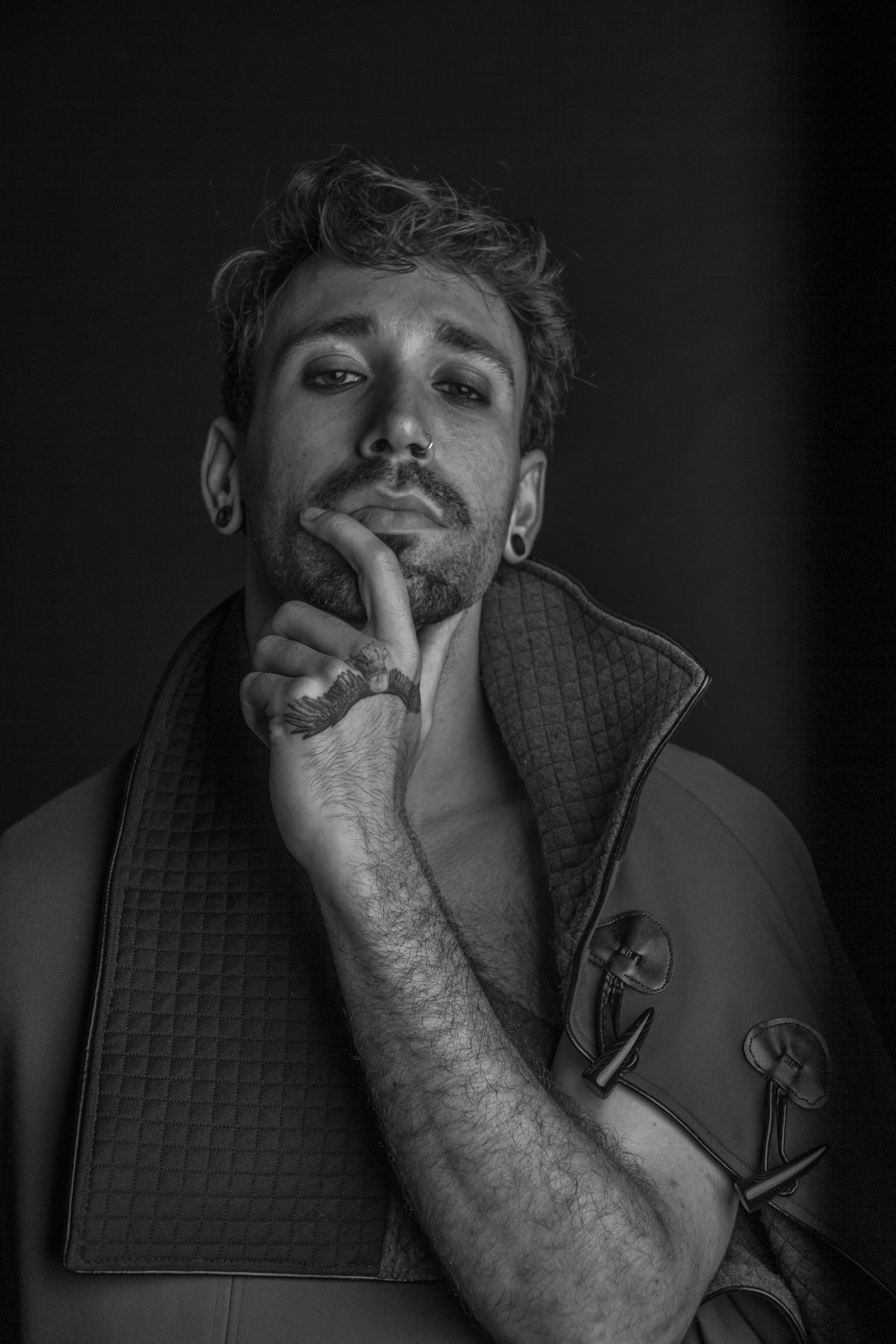 Black and white portrait of a young man with tattoos, piercings, and styled hair, wearing a jacket, posing with his hand resting on his chin.
