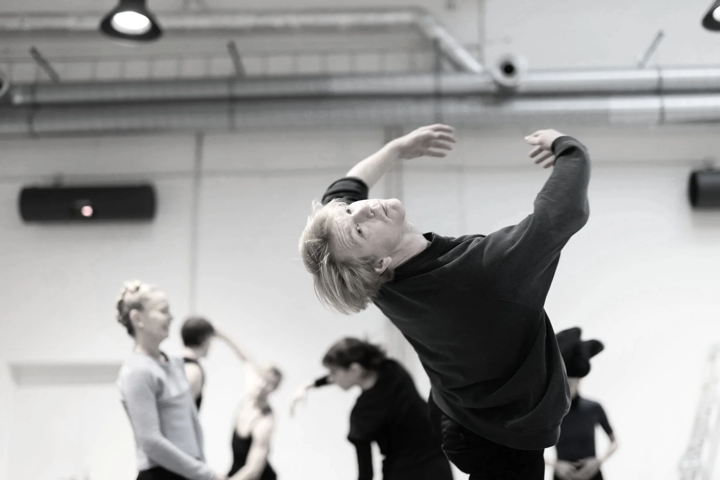 A person with blond hair stretching with arms raised in a dance or yoga studio, with several other people in the background practicing similar movements.
