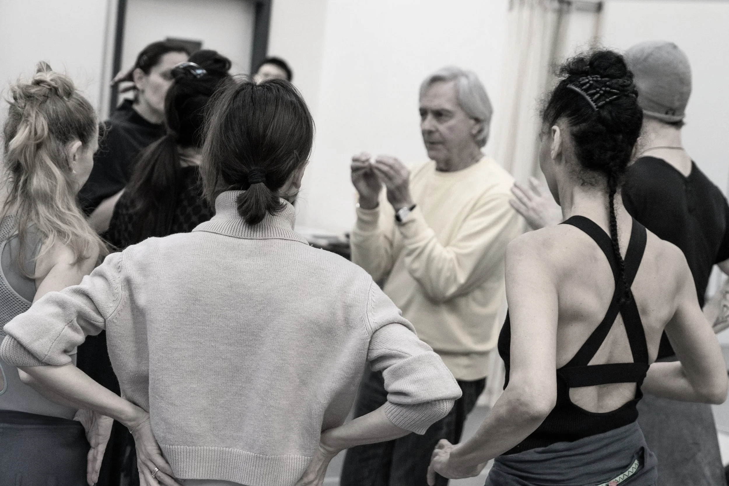 A group of people gathered around a man who appears to be giving instructions or a talk, in a casual setting. The group includes women with diverse hairstyles and a man in a hat, and they are listening attentively.