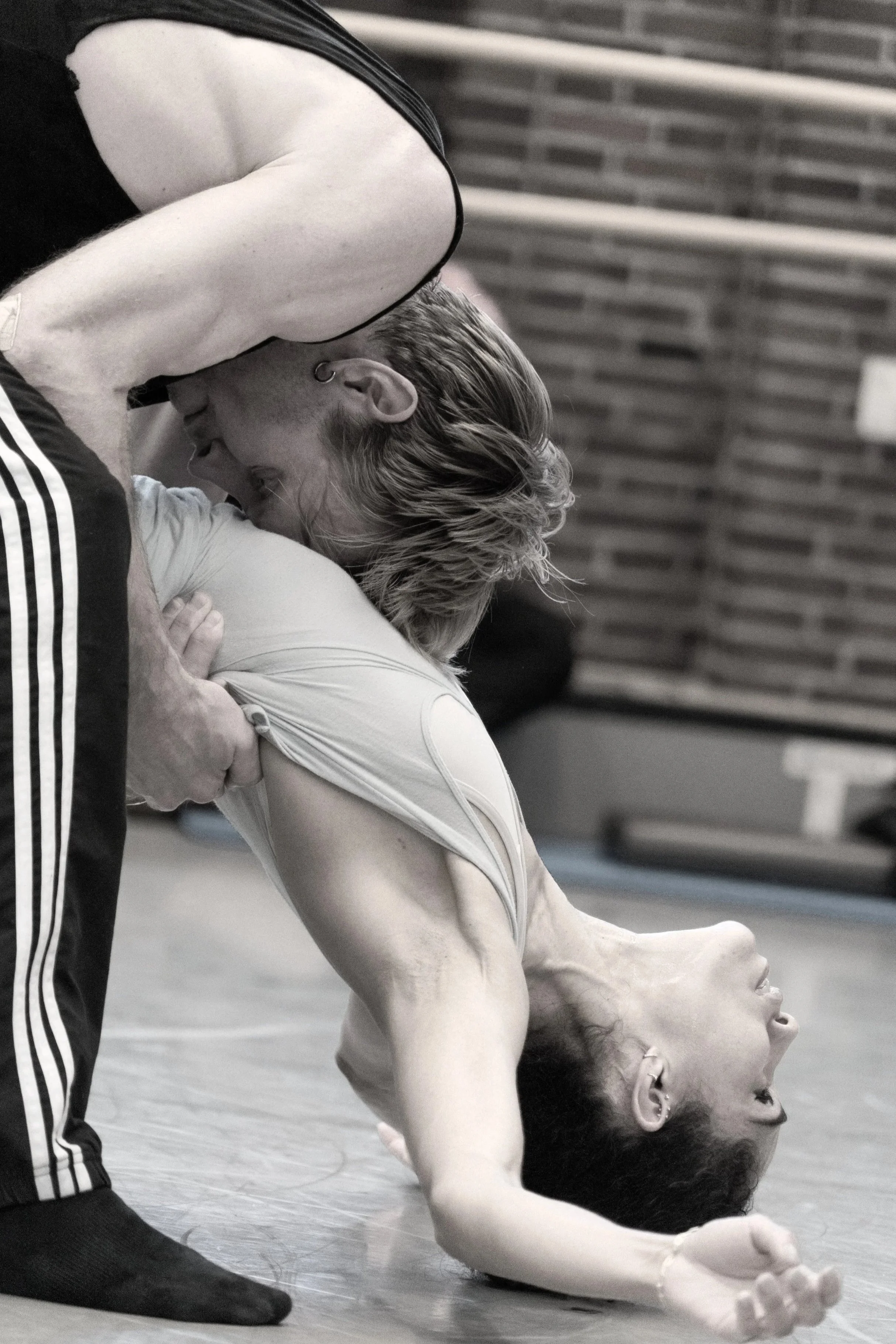 Two dancers perform an acro dance move on the floor, with one dancer lying on her back and the other leaning over her, touching foreheads. The image is in black and white.