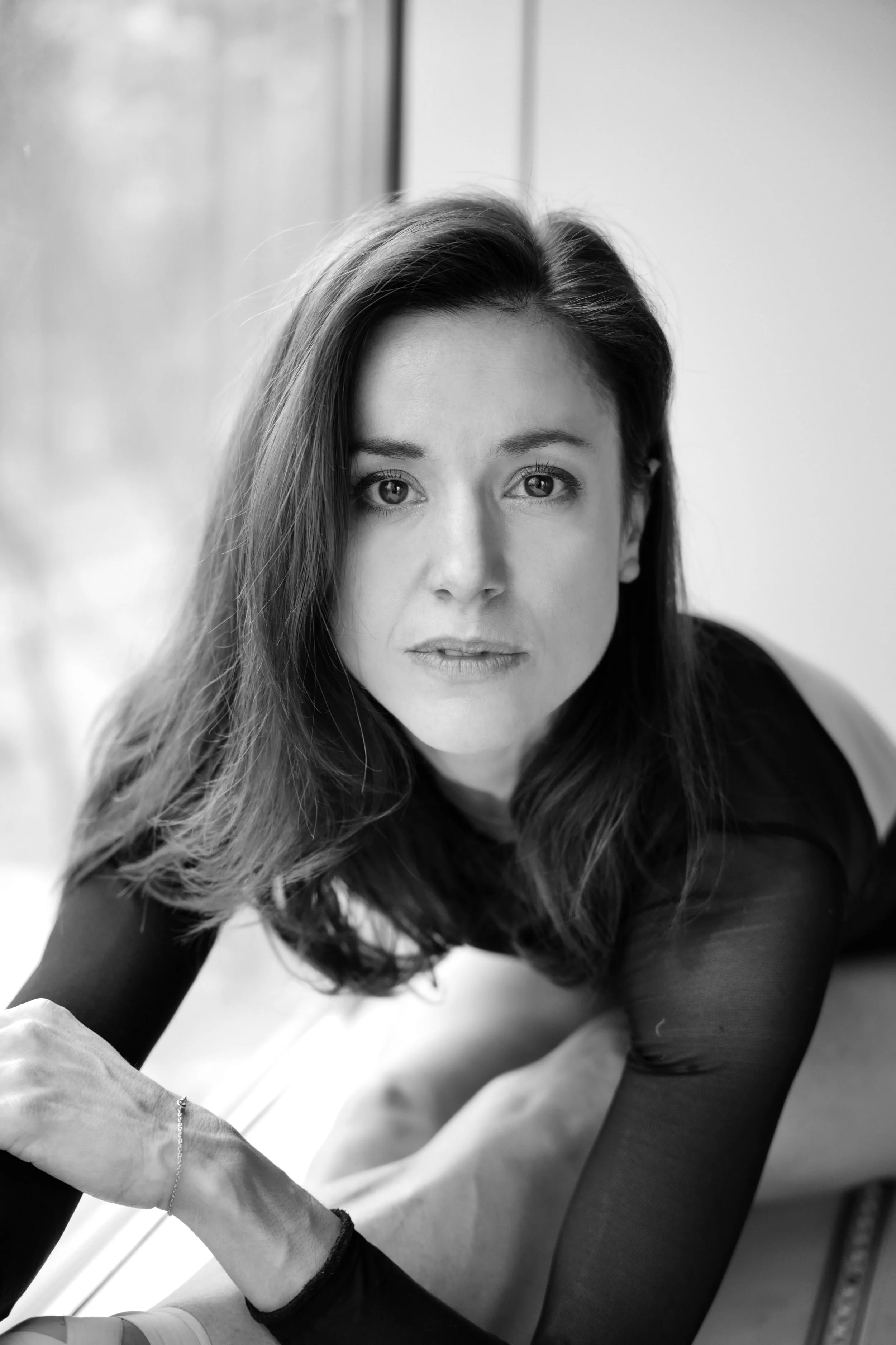 Black and white portrait of a woman with long dark hair, looking directly at the camera with a serious expression, leaning on a surface.