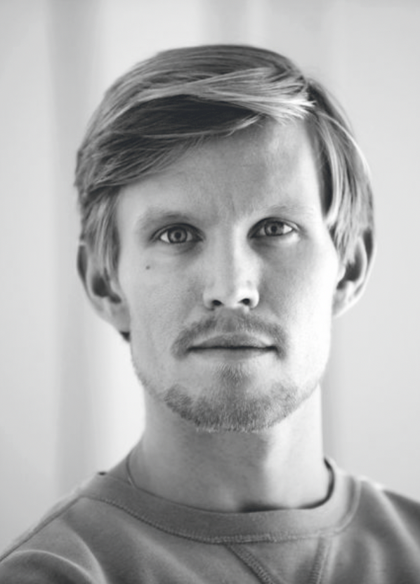 Black and white portrait of a young man with medium-length hair, light facial hair, and a neutral expression, looking directly at the camera.