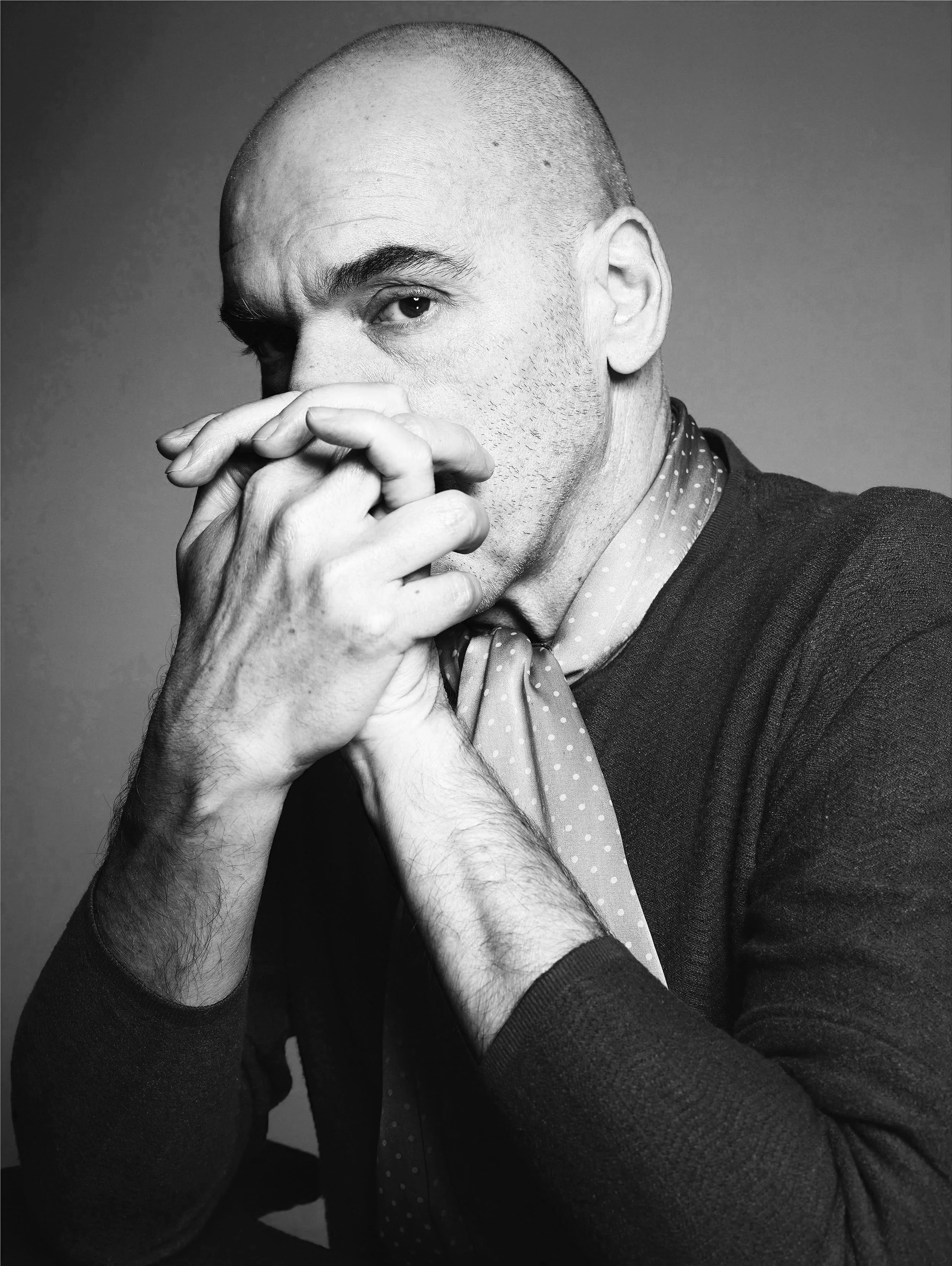 Black and white portrait of a bald man with his hands covering his mouth, wearing a dotted scarf and dark clothing, looking intently at the camera.