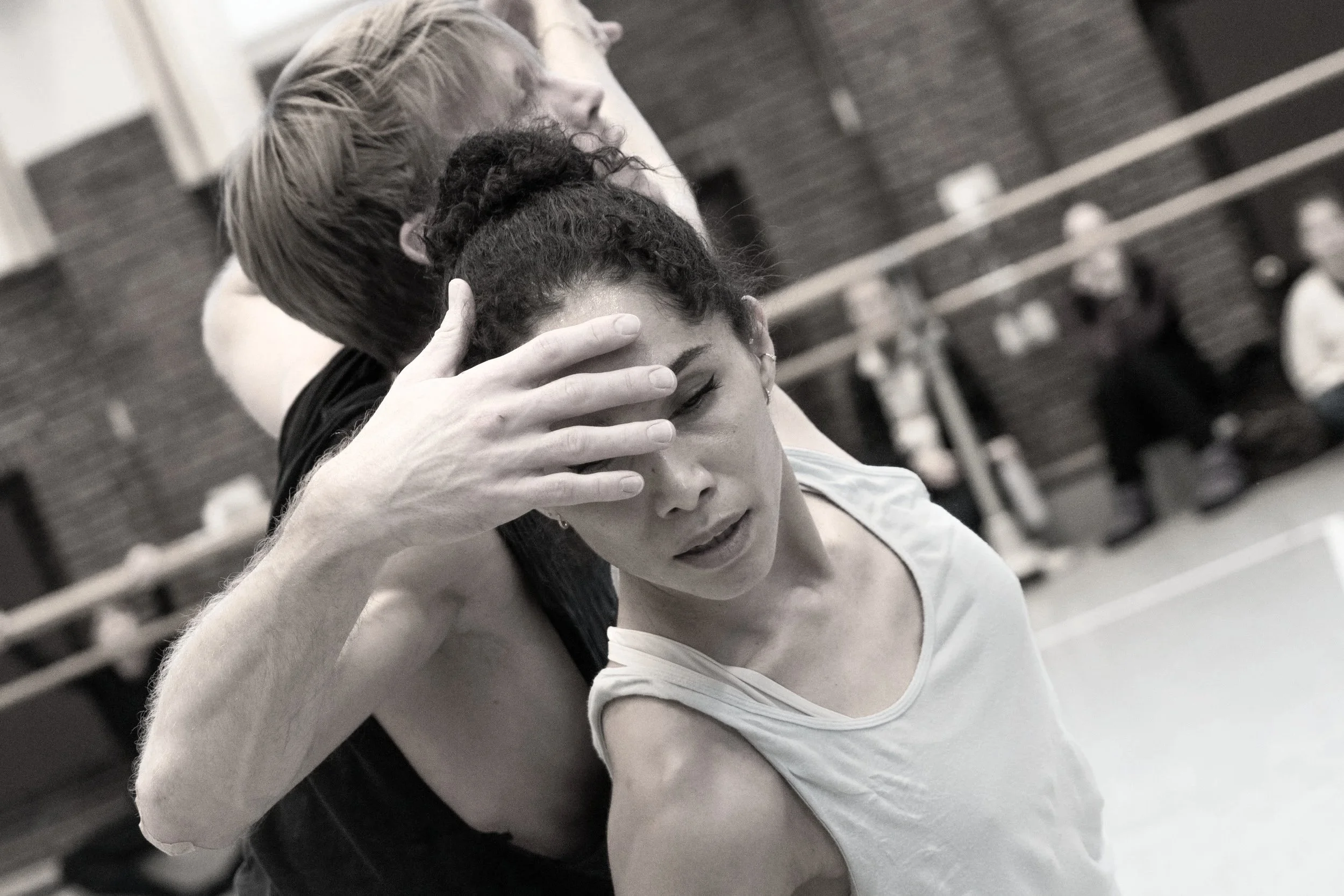 A dance instructor supports a young female dancer during a practice session in a gym, holding her head gently and guiding her as she appears to be emotional or tired.