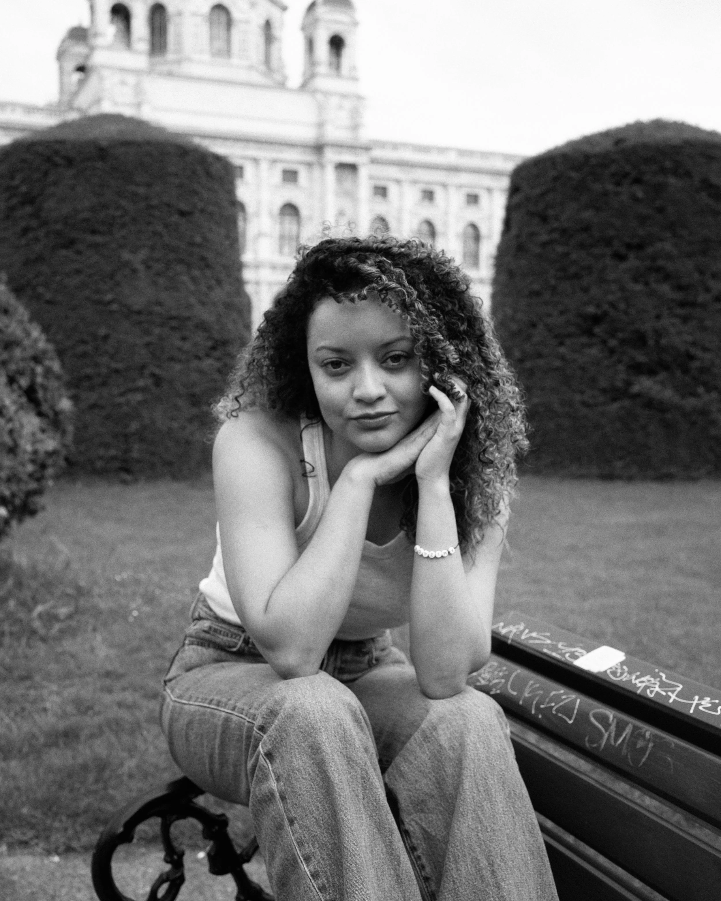 A woman with curly hair sitting on a park bench, resting her chin on her hands, with a large historical building in the background.